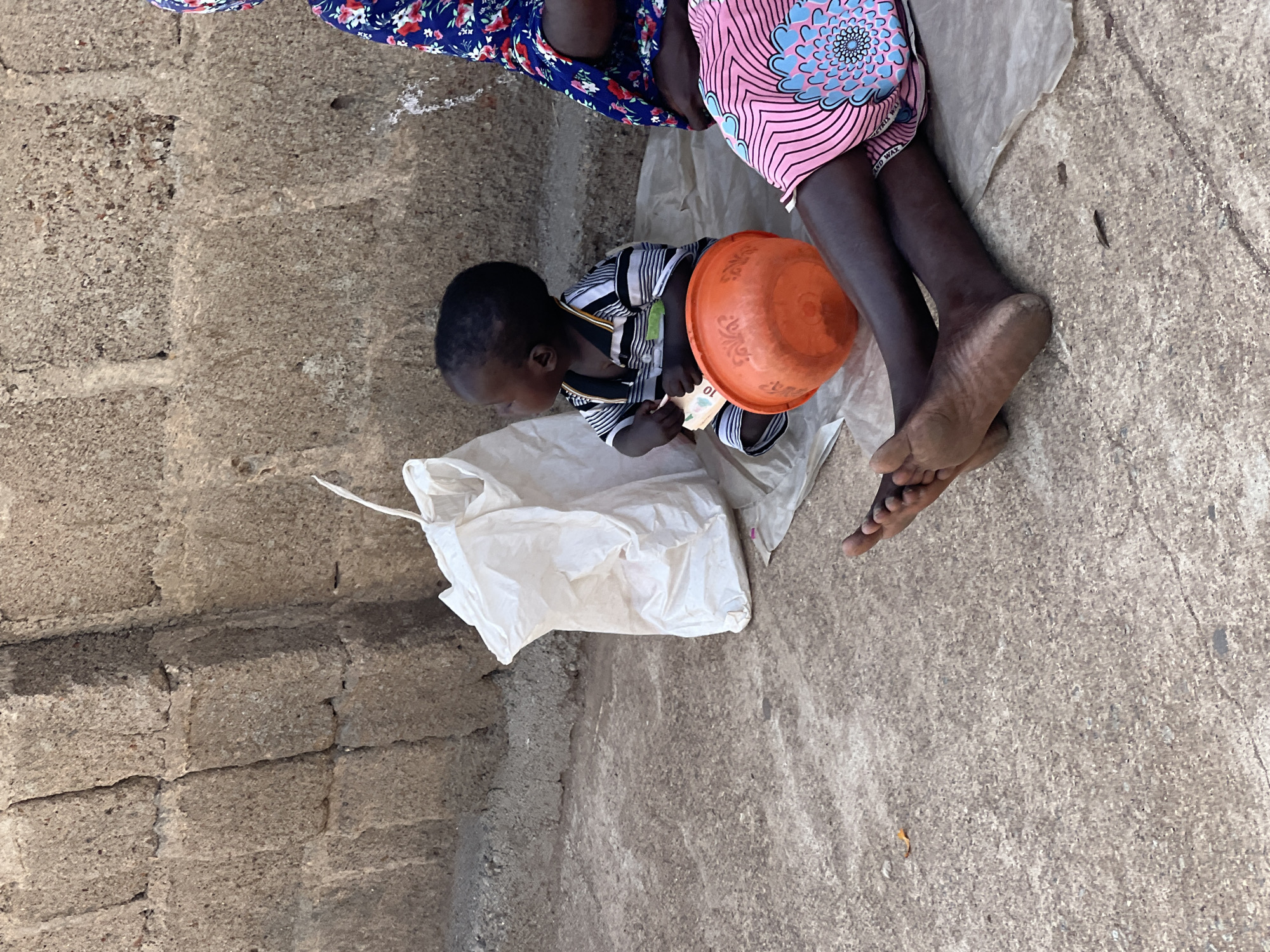 A young Black child in a black and white striped shirt sits on a sheet of paper on the ground next to an orange bowl, holding a bill. An adult's bare foot is next to the child. Two adults are sitting close by, one wearing a floral dress, the other wearing a pink patterned dress. The setting appears to be outdoors on a paved or concrete surface, possibly near a brick wall. This photograph was taken in Jos, Nigeria.
