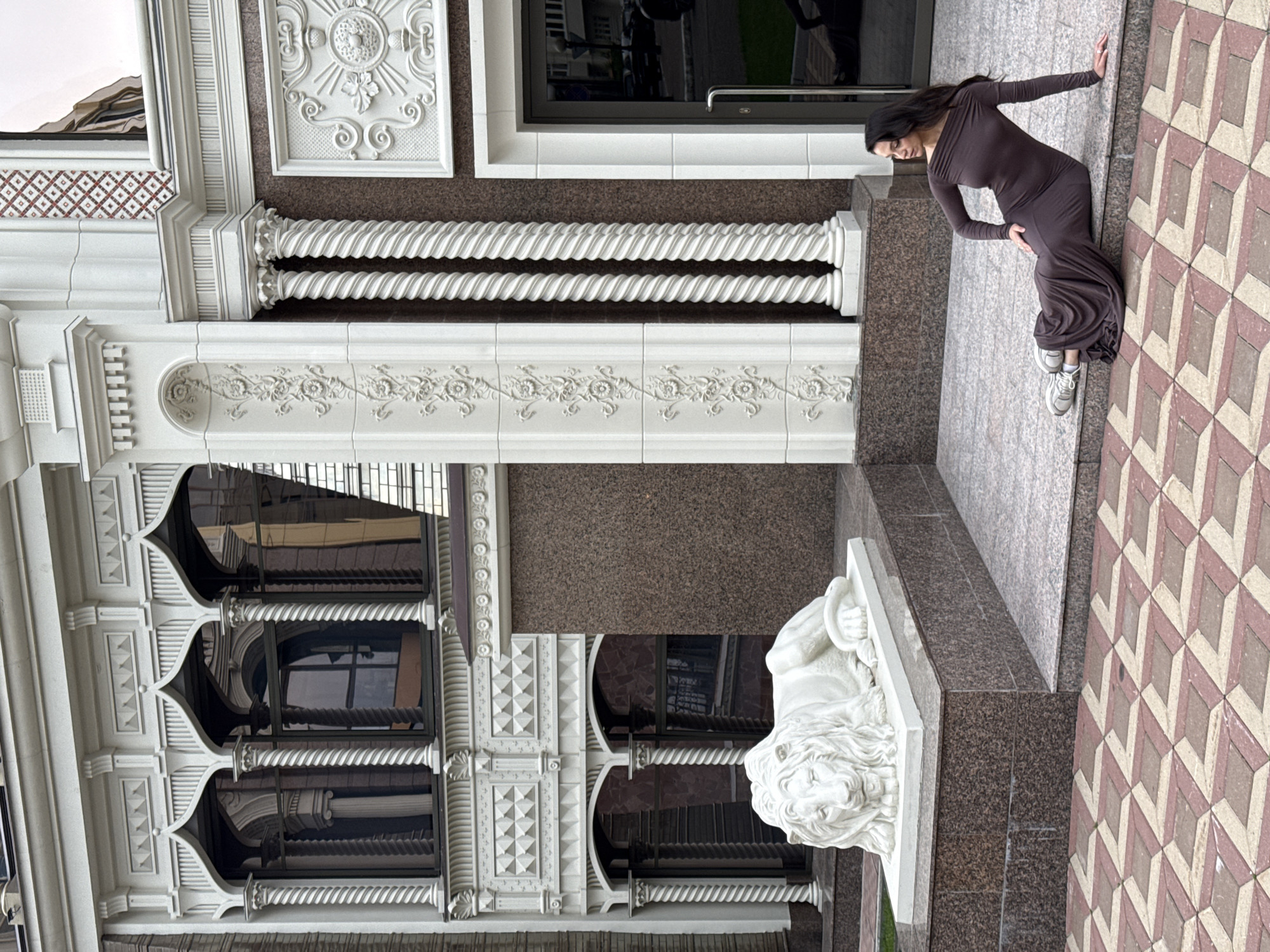 The image captures an ornate building with a neo-Moorish architectural style, featuring arched windows with decorative columns and elaborate white stucco detailing. In the foreground, a white lion sculpture rests on a tiered granite base. To the right of the sculpture, a woman with long dark hair is seated on the steps leading up to the building. She is wearing a long, dark brown, off-the-shoulder dress and grey sneakers. The woman is looking to her left, away from the camera, with her right hand resting on her hip and her left arm extended towards the ground. The ground in front of the steps is paved with a geometric pattern of red and beige tiles. The building appears to be a private residence or a commercial establishment, possibly located in Kazan, Russia, given the architectural style which can be found in some affluent areas of the city.