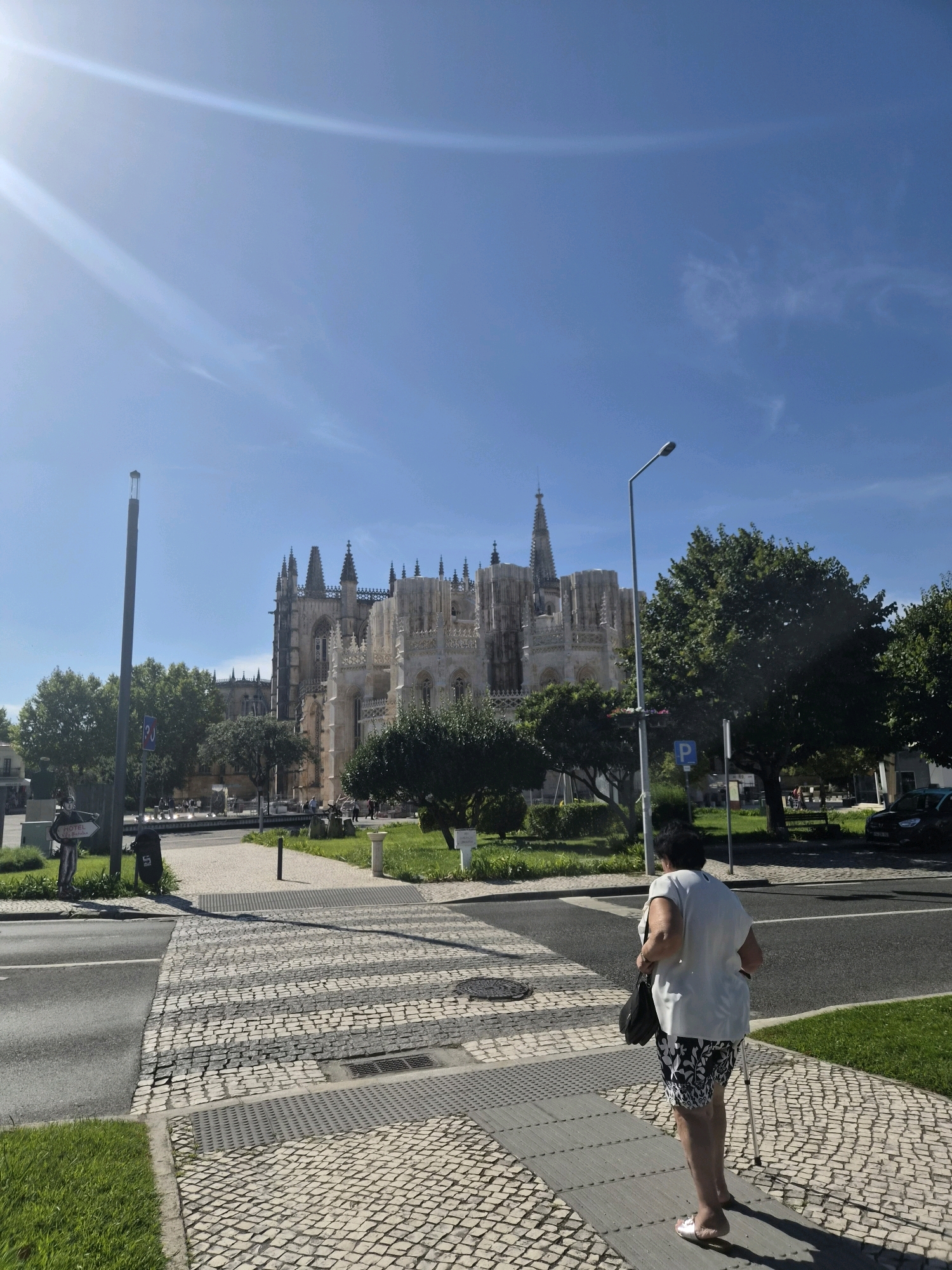 An overhead shot captures a sunny day in Batalha, Portugal, near the Monastery of Batalha. The monastery, a grand Gothic structure with intricate stonework and spires, dominates the background under a clear blue sky with sun flares. In the foreground, a woman with dark hair and a white top walks away from the camera, holding a black purse and using a cane. She wears a patterned skirt and sandals. She is on a sidewalk paved with cobblestones, which transitions into a crosswalk made of similar stones. To the left of the sidewalk, a grassy area with trees and bushes leads to a street with parked cars and a lamppost. A street sign and a statue of a person holding a sign are also visible on the left side of the image. The scene depicts a moment of everyday life against the backdrop of a significant historical landmark.