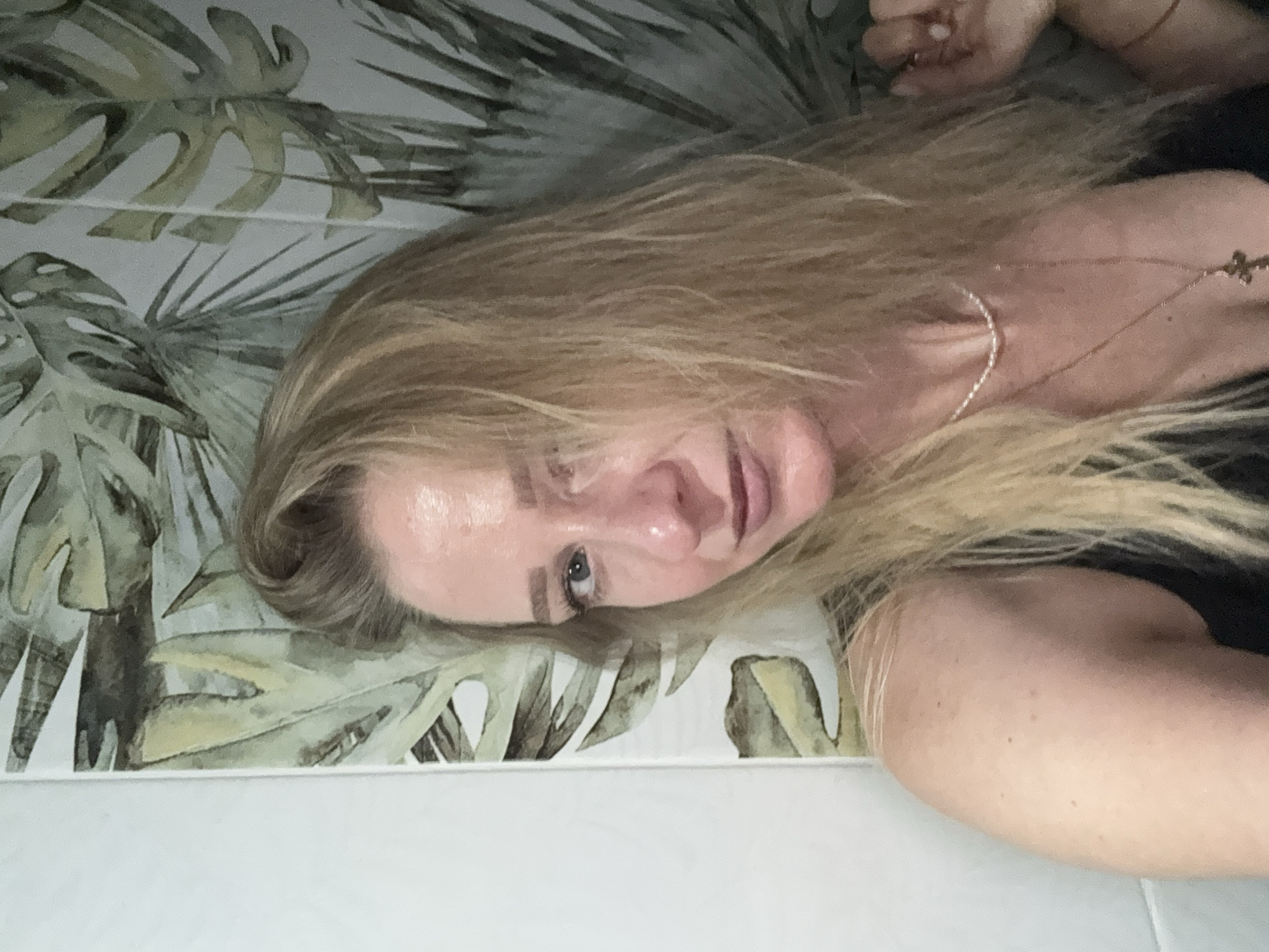 A fair-skinned blonde woman is in a bathroom with a tropical leaf-patterned wallpaper. She is looking at the camera, and her right hand is partially visible at the right edge of the frame. She is wearing a black tank top and a pearl necklace with a cross pendant. The lighting appears to be natural, as evidenced by the highlights on her forehead and nose.