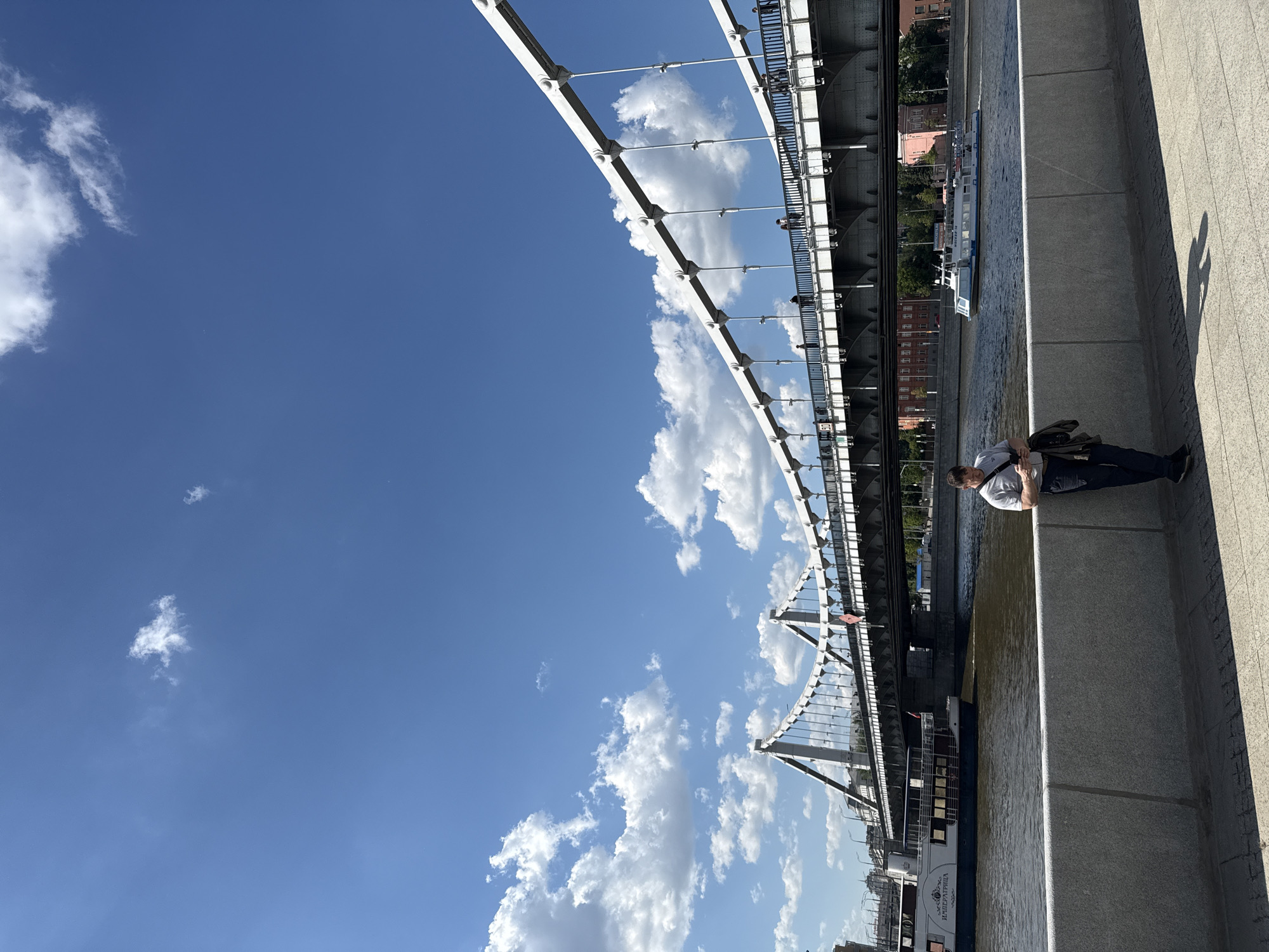The image depicts a bright, sunny day along a river in Moscow, featuring a prominent bridge, a river cruise boat, and a man in the foreground. The sky is a clear, vibrant blue with scattered white cumulus clouds, indicating fair weather.
In the foreground, on the right side of the frame, a man wearing a light-colored t-shirt and dark pants leans casually against a tall, grey concrete embankment. He appears to be looking down, possibly at his hands or a mobile device. His shadow is cast sharply on the pavement behind him, suggesting midday or early afternoon sunlight.
To the left of the concrete wall, a wide river flows. A large passenger boat, possibly a river cruise ship, with a white upper deck and a dark hull, navigates the water. Clearly visible on its side is the Cyrillic text "ИМПЕРИАЛ РИВЕР" (Imperial River).
Dominating the mid-ground and left side of the image is a large suspension or cable-stayed bridge, characterized by its white structural elements and suspension cables. People can be seen walking along the bridge's pedestrian pathway in the distance. Beyond the river and under the far side of the bridge, several buildings with reddish-brown facades and green trees are visible on the distant bank. The overall scene captures a serene urban landscape under clear skies.