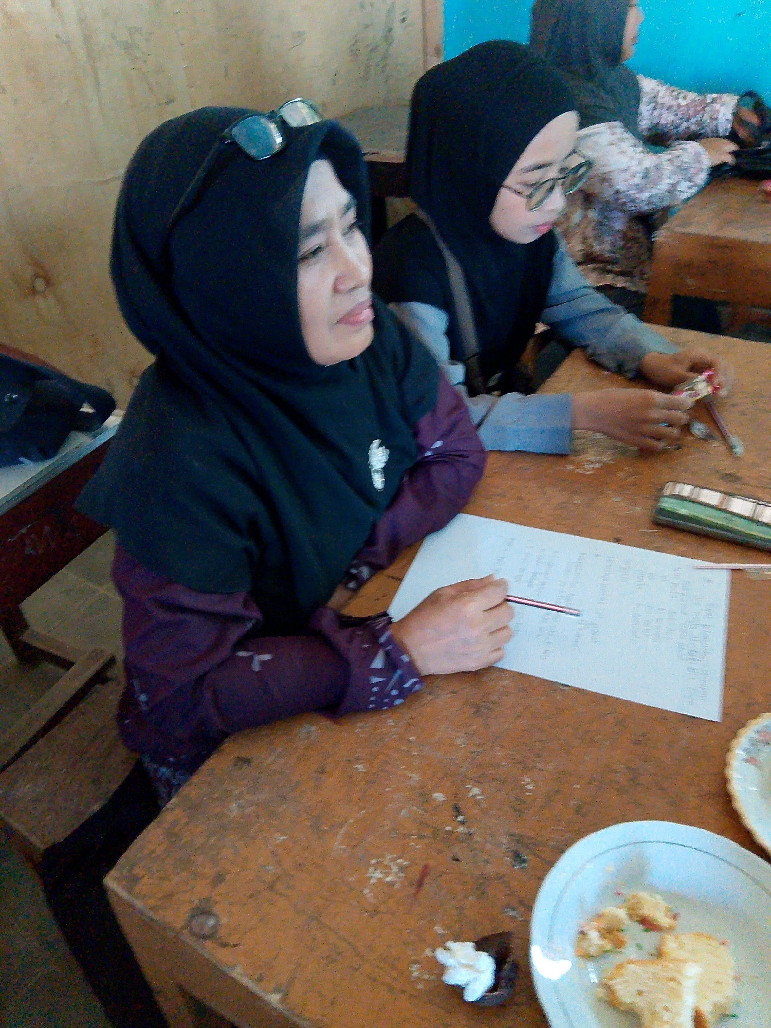 This image depicts an indoor scene, likely a classroom or a community gathering space, featuring two young women wearing hijabs. The woman in the foreground, positioned on the left, is wearing a black hijab and a dark purple, patterned long-sleeved shirt. She is looking to her right and has a pair of glasses resting on top of her hijab. Her right hand is holding a pen over a piece of paper with handwritten text. Beside her, on the right, is another young woman wearing a black hijab and a grey and black long-sleeved shirt. She is looking down at her hands and appears to be interacting with some small objects, possibly a pencil sharpener or craft supplies.
The environment is characterized by wooden tables. The table in the foreground is a light brown, worn wood surface. There are several items on the table, including a white plate with pieces of cake or pastry and some colorful sprinkles, and a small dark container with a white dollop of frosting. A small pencil case or container with stripes is also visible on the table. The background shows a textured, light-colored wall. The overall lighting suggests an indoor setting, possibly with natural light filtering in from an unseen source. The image appears to be taken in Kuripan Utara, Indonesia, though specific landmarks or contextual clues within the image to confirm this location are not readily apparent.