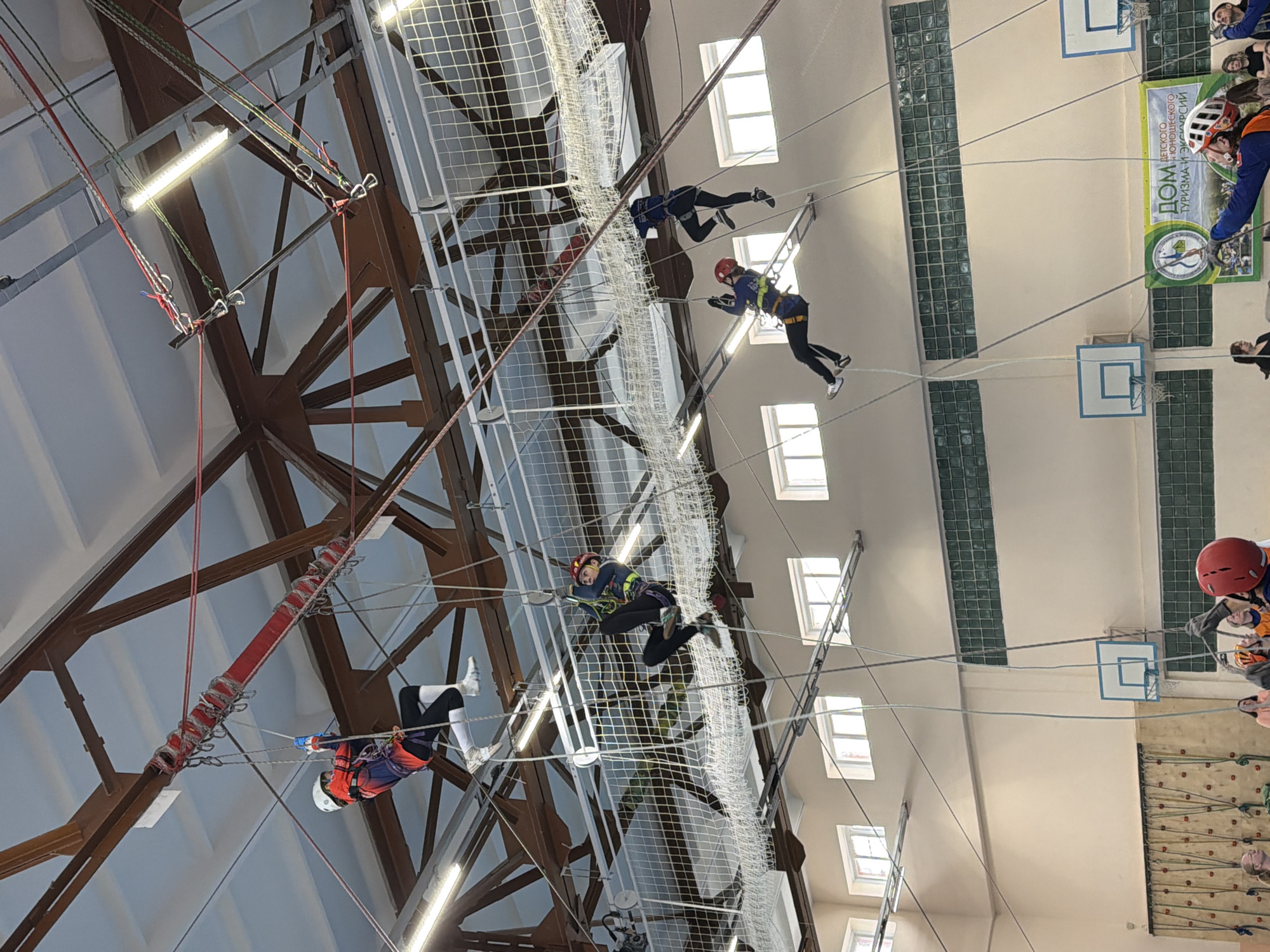 The image depicts an indoor ropes course, likely a recreational or training facility. Several individuals are navigating the course, attached to safety harnesses and ropes. The course includes rope bridges, climbing nets, and suspended obstacles. The building appears to be a gymnasium or large indoor space with a high ceiling supported by metal beams. Windows line the walls, providing natural light. A sign in Russian is visible, suggesting the location is in a Russian-speaking area. Based on the provided context, this activity is occurring in the city of Berezniki, Russia.