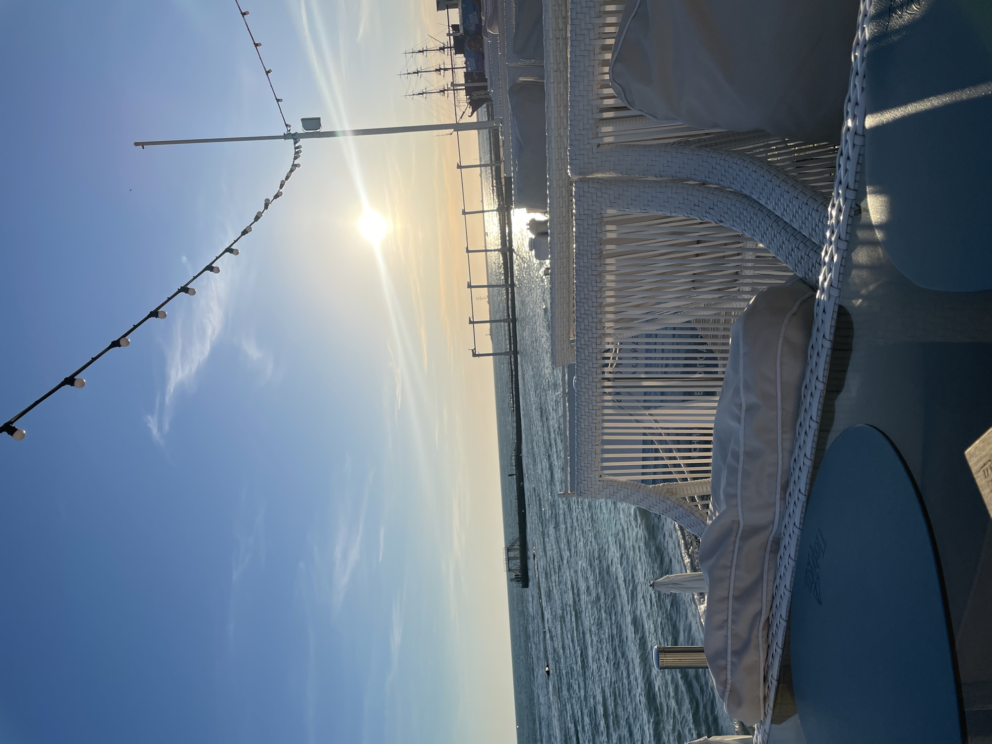 The photograph depicts a seaside cafe with white wicker furniture and cushions, overlooking the Black Sea at sunset. The sun is low in the sky, casting a golden glow on the water and creating reflections. In the distance, a pier extends into the sea, and a tall ship with sails is docked. String lights are hung overhead, adding to the ambiance of the scene. The location is likely a cafe or restaurant along the coast of Sochi, Russia, known for its beachfront establishments and scenic views. The image captures a serene and picturesque moment, evoking a sense of relaxation and enjoyment.