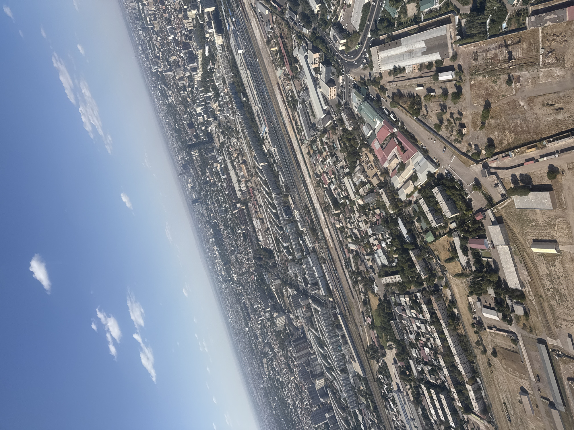 This is an aerial view of a densely populated urban area in Tashkent, Uzbekistan, under a clear blue sky with scattered white clouds. The perspective is from a high angle, looking down and slightly to the side, revealing a vast expanse of buildings, roads, and industrial infrastructure.
In the foreground, there are large, industrial-looking buildings with metal roofs, some appearing to be warehouses or factories, situated on barren, dusty land. Adjacent to these are areas with smaller residential buildings, characterized by flat roofs and various colors, interspersed with trees. A prominent road with several vehicles, including a bus, winds through this section of the city.
Further into the midground and background, the cityscape unfolds into a mosaic of multi-story residential buildings, typical of Soviet-era architecture, with long, uniform facades. These are laid out in rectangular blocks, with green spaces and trees visible between them. A significant railway line with multiple tracks runs horizontally across a large portion of the image, passing by industrial buildings and residential areas, suggesting it is a major transportation artery. Beyond this, the city stretches towards the horizon, with a dense concentration of buildings, some of which are taller modern structures, indicating a mixed architectural landscape. The overall impression is of a bustling city with a clear division between residential, industrial, and commercial zones, all interconnected by a network of roads and railways.