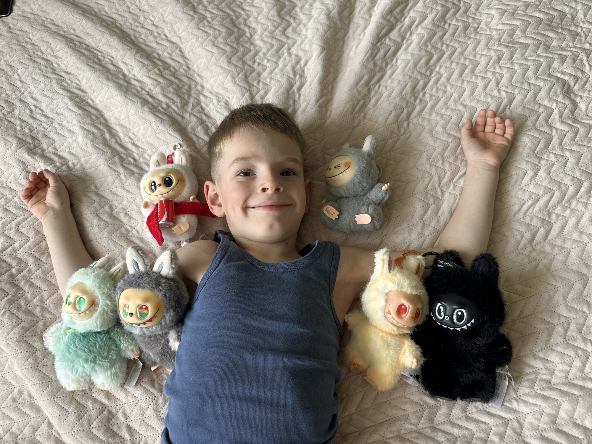A young boy is lying on his back on a bed with a light beige patterned quilt in Chaikovsky, Russia. He wears a navy blue tank top. He is smiling and has his arms outstretched on either side of him. He is surrounded by six plush toys: two are grey and white rabbits with small, dark eyes and red scarves, one is grey with small pink toes and a smiling face, two are a light green and grey rabbit with a distinct doll-like face, and one is a black rabbit with a smiling face. The black rabbit has tags attached.