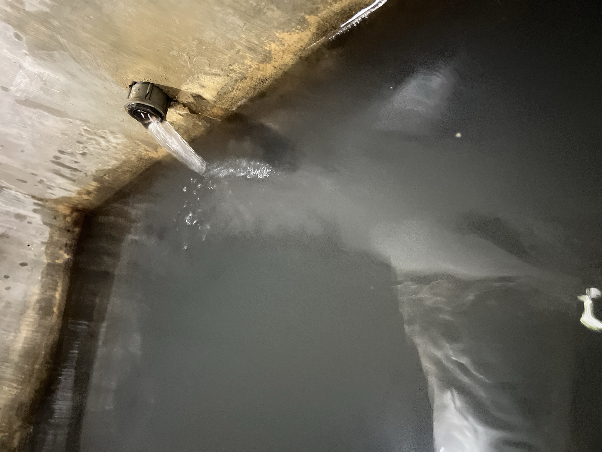 In Kilometer Delapan, Indonesia, a concrete cistern contains dark water. A pipe protruding from the cistern wall is actively discharging a stream of clear water, creating ripples and mist on the surface of the dark water. The concrete cistern appears aged and stained, with the water level nearing the top edge. The surrounding environment is not visible.