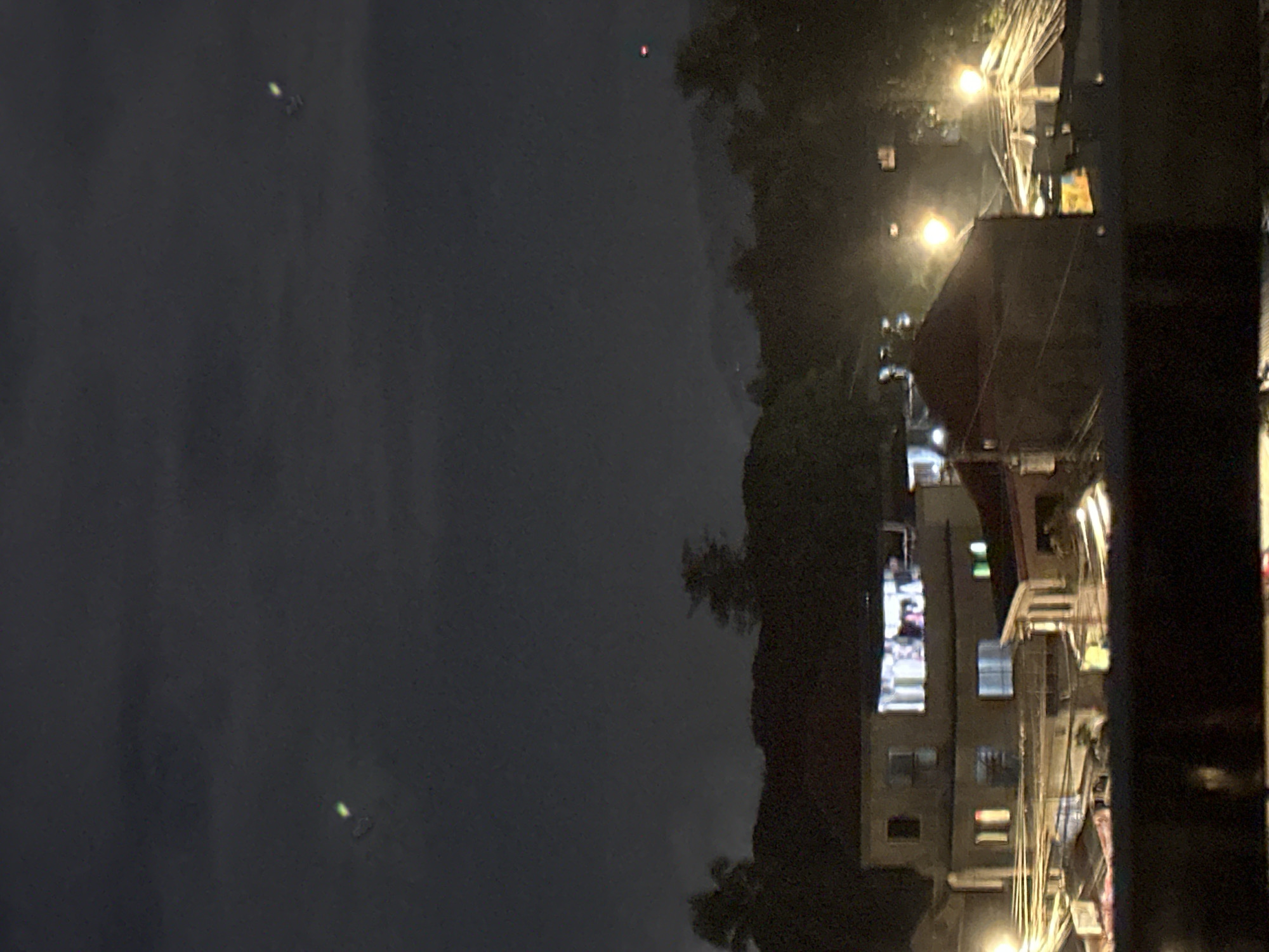 The image shows a nighttime view of buildings and trees in Davao City, Philippines. There are lights visible from windows and streetlights. In the sky, there are three blurry objects that appear to be lights. One is a greenish-white streak in the upper left, another is a greenish-white object in the upper right, and a third is a reddish-orange object in the upper right. The scene depicts a residential or commercial area at night.