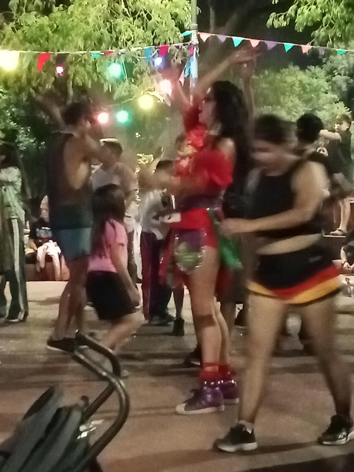A vibrant street scene unfolds with people gathered for a celebration. A woman stands out in a red, ornate outfit and purple sneakers, her arms raised joyfully. Colorful pennant banners and string lights hang overhead, adding to the festive atmosphere. Other figures are blurred, suggesting movement and activity. A stroller handle is visible in the foreground. A girl kneels on the left, possibly observing the spectacle.