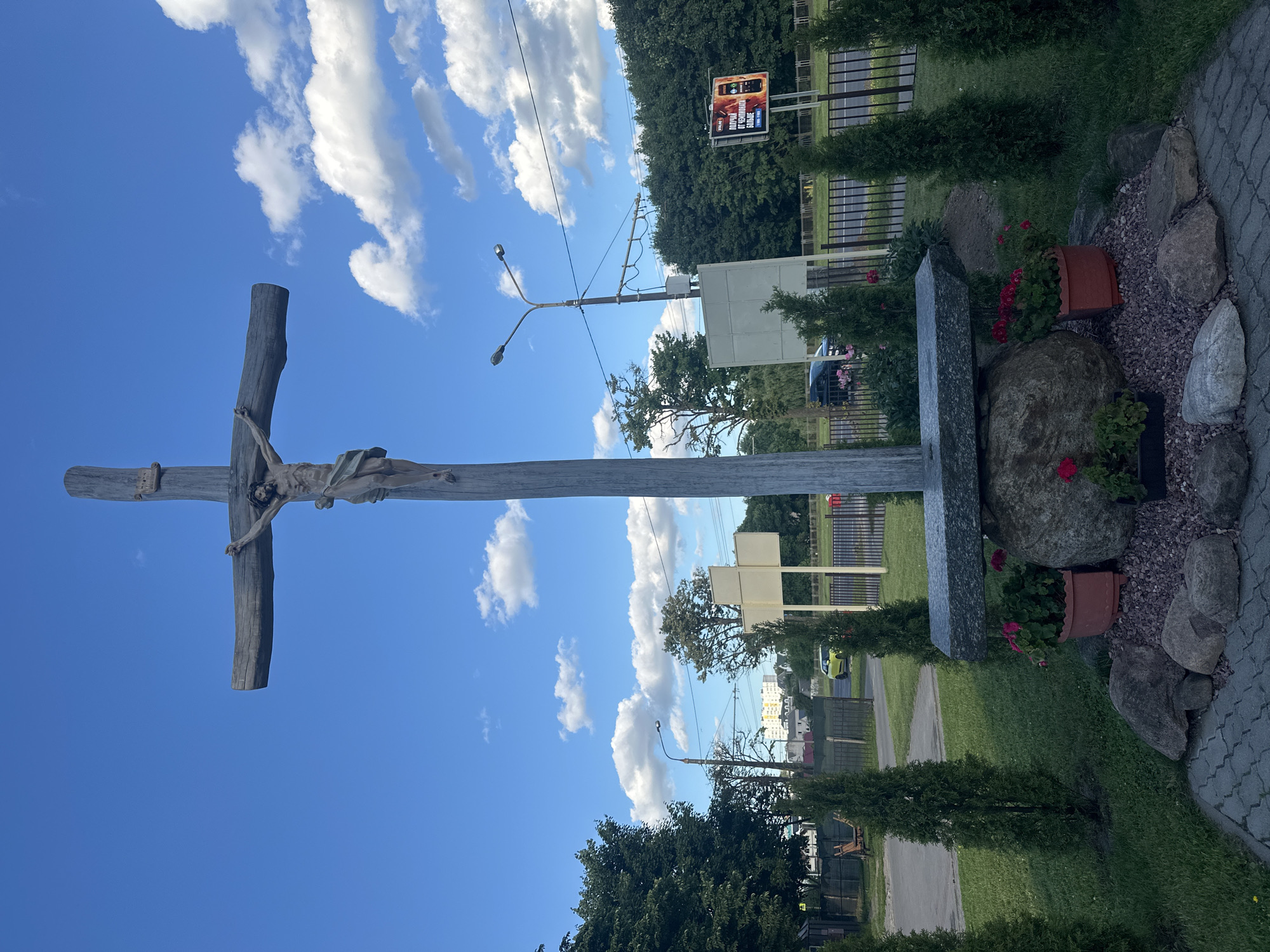 The image depicts a wooden crucifix with a sculpture of Jesus Christ against a bright blue sky filled with fluffy white clouds. The crucifix is large and appears weathered. To the right of the crucifix, there is a stone monument, rocks, and potted plants with red flowers. The scene is outdoors, with a paved walkway and grassy areas. In the background, there are trees, a fence, streetlights, and a billboard displaying an advertisement with text in Cyrillic script. Further in the background, buildings and a street with a parked car are visible. The lighting suggests it is daytime, likely a sunny day with partly cloudy weather. The overall mood is solemn due to the religious imagery. The location context is Minsk, Belarus.
