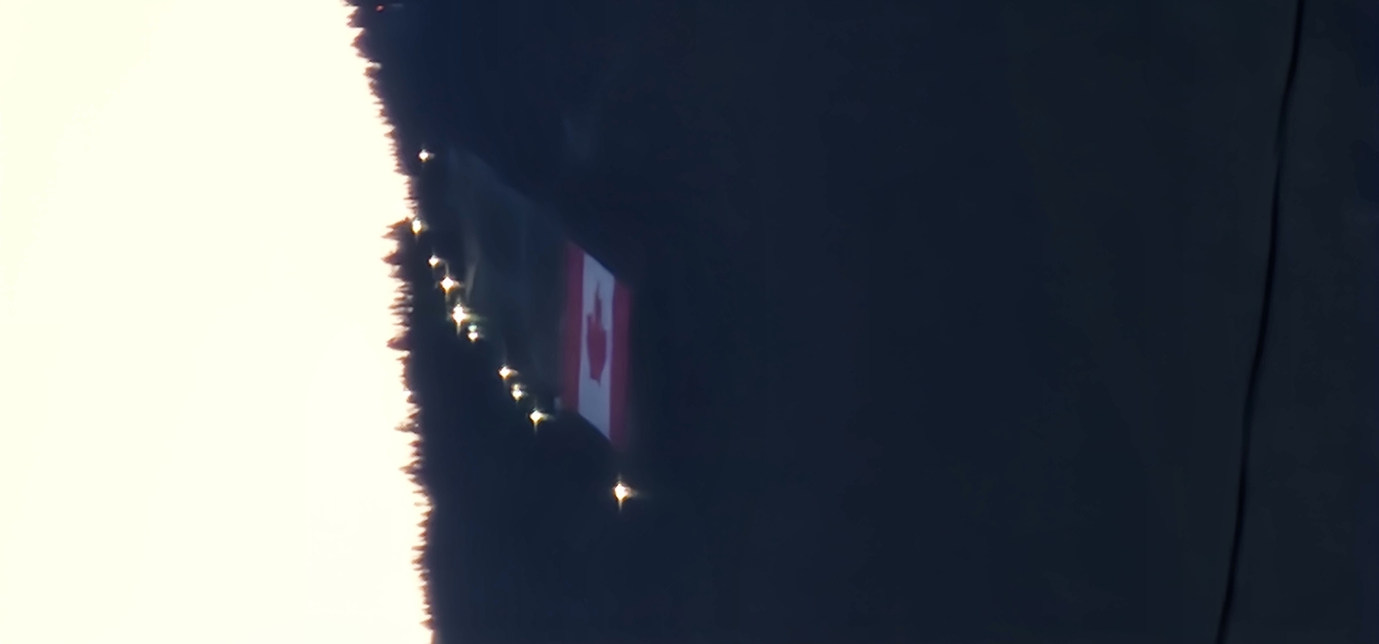A wide shot captures a Canadian flag illuminated on a dark, silhouetted hillside at dusk. The flag, a prominent red and white maple leaf design, is positioned on the slope of a hill lined with trees, also dark silhouettes against the pale sky. A row of bright, white lights is visible ascending the hill, suggesting a path or ski slope. The overall scene is dark, with the exception of the illuminated flag and lights, and the sky is a soft, bright gradient. The image does not show any people, city buildings, or other specific identifiable landmarks, making it impossible to determine a precise location within a city.