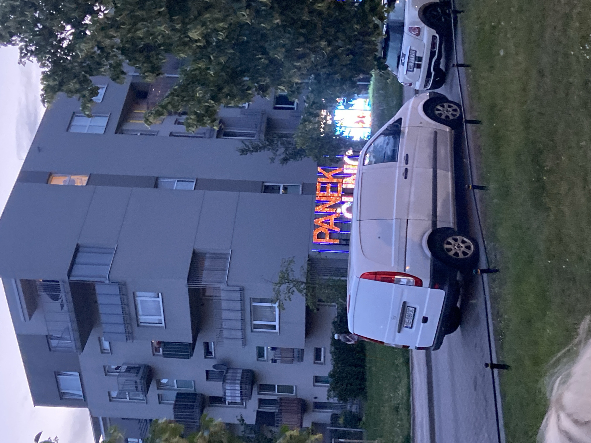 In Warsaw, Poland, an urban scene unfolds at dusk. A modern, grey apartment building dominates the background. Numerous balconies with metallic railings jut out from its facade, and windows show signs of internal lighting. In the middle ground, a neon sign displays "PANEK" in large, illuminated letters, with more colorful, abstract neon lights below. To the left, a white van with its rear doors open is parked on a paved surface adjacent to a grassy area with a black decorative fence. To the right, a white Mini Cooper is parked. A solitary figure, partially obscured, stands near the van. Lush green trees frame the right side of the image. The sky is overcast, indicative of the evening hour.