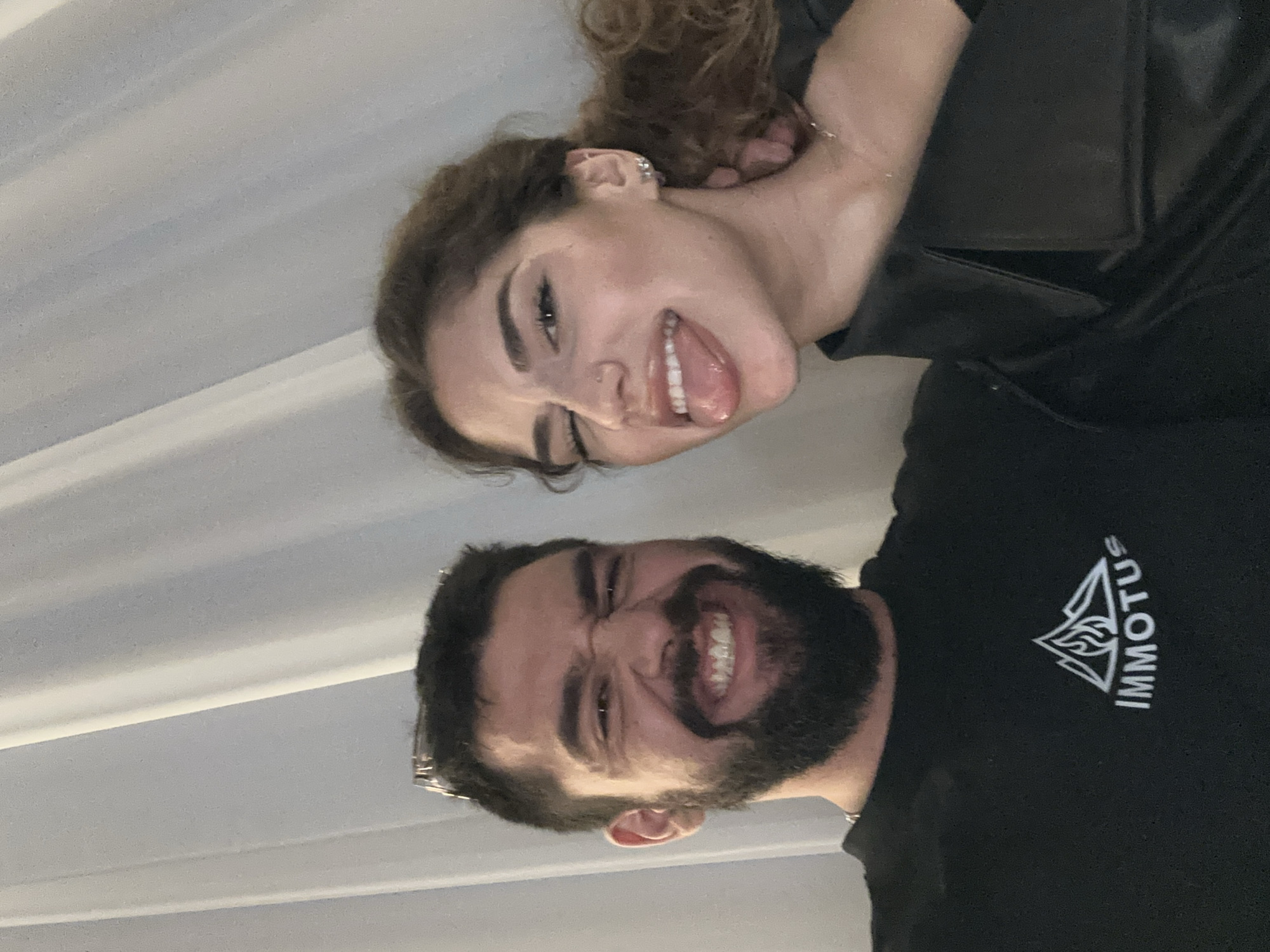 Two individuals are positioned closely together in a photograph. A woman with curly brown hair, wearing a black leather jacket, is at the top of the frame sticking out her tongue. Below her is a man with a full dark beard, wearing a black t-shirt with the word "IMMUTUS" and a logo printed on it. The background is a plain white surface. The location is unknown, and there is nothing visible in the photo to indicate that it was taken in Plovdiv, Bulgaria.