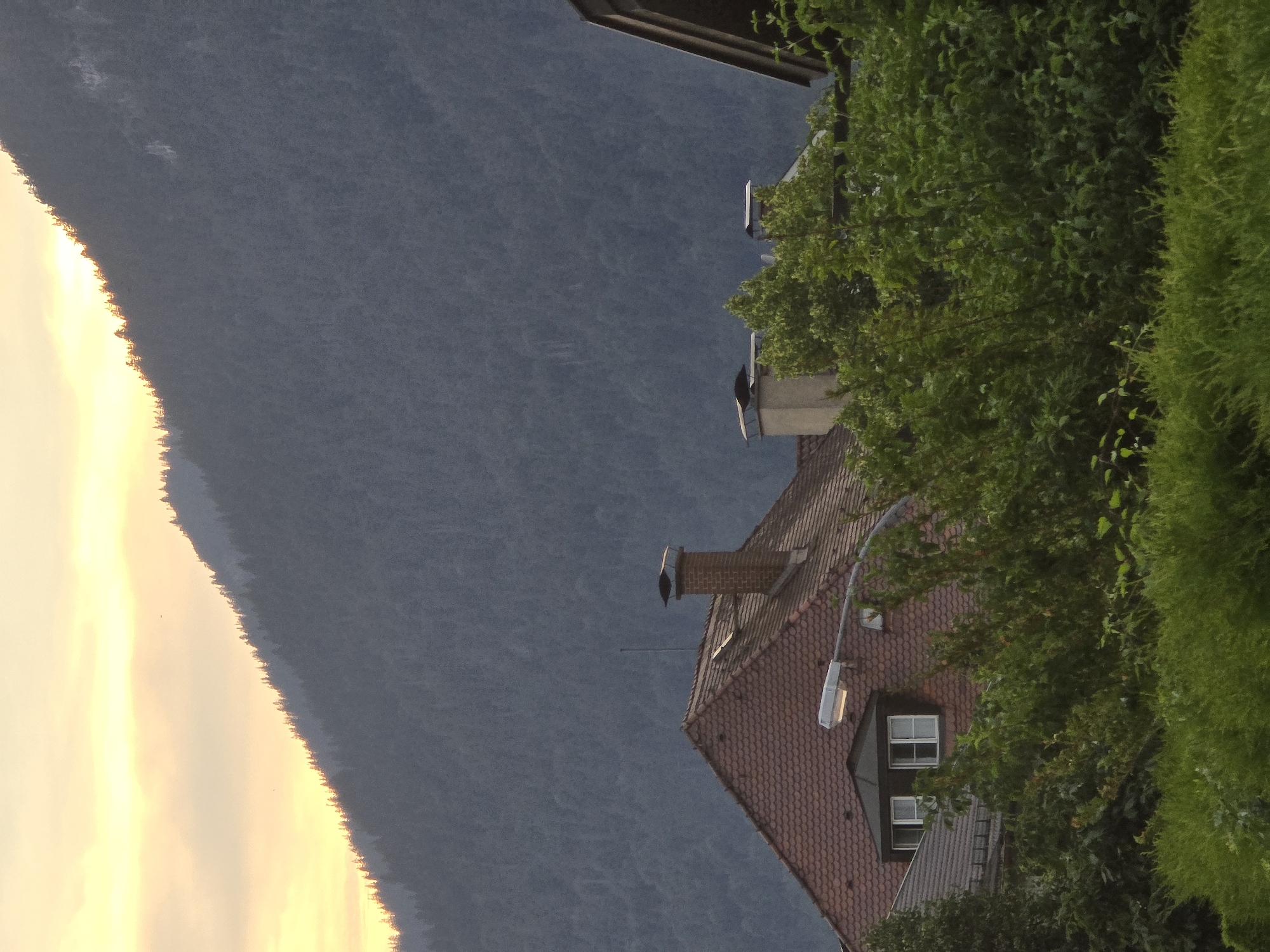 This is a photograph of a mountainside with a small village in the foreground. The mountain is covered in dark green trees, and the sky is a pale yellow with some clouds. In the foreground, there are several houses with red tiled roofs. One house has a red brick chimney. There is also a street lamp and some trees. The scene appears to be set in the late afternoon or early evening, as the sun is low in the sky. The image was taken in Garmisch-Partenkirchen, Germany.