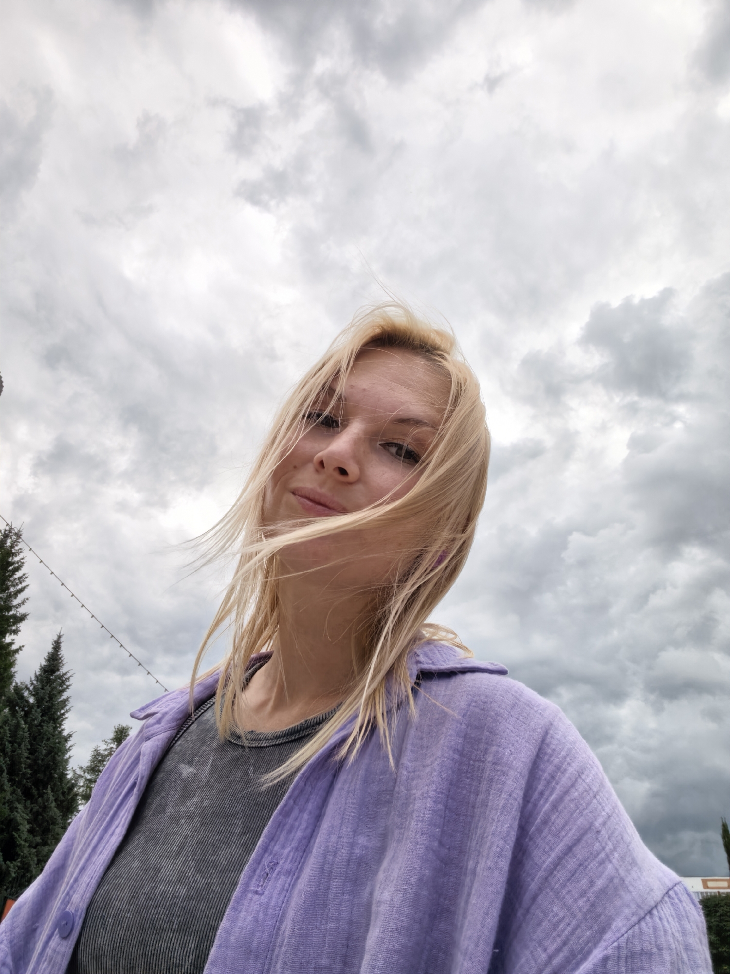 A young woman with blonde hair, wearing a grey t-shirt and a purple open shirt, is captured in a low-angle selfie against a cloudy sky. Her hair is blowing in the wind, framing her face. In the background, evergreen trees with string lights are visible on the left side of the frame, suggesting an outdoor setting, possibly a park or a street with decorations in Sterlitamak, Russia.