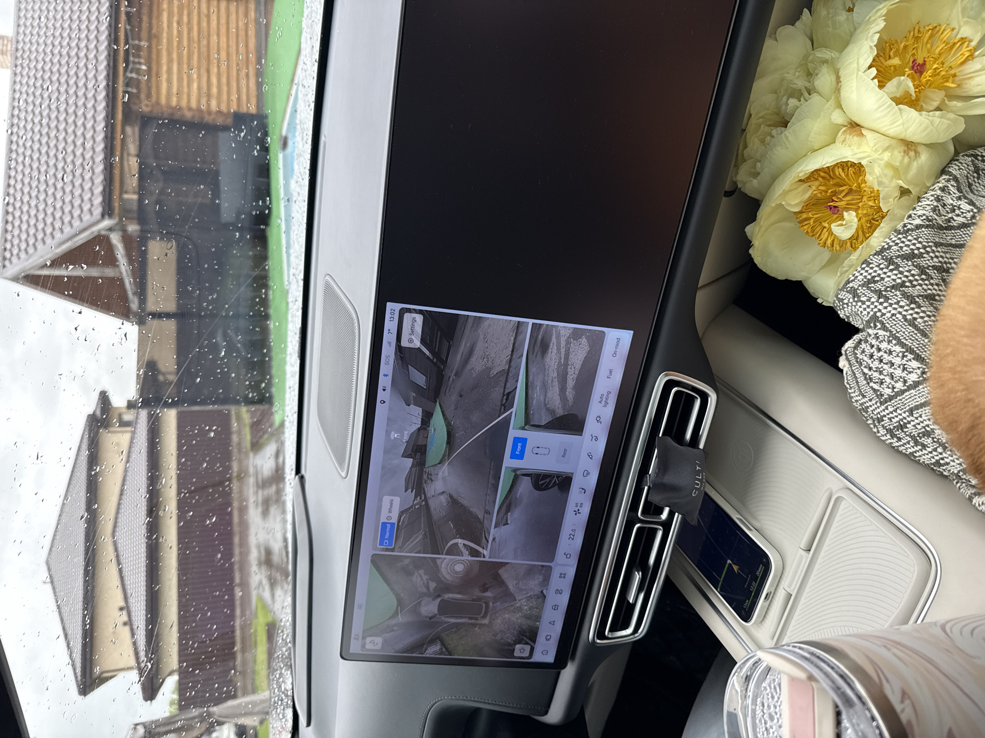 The image shows the interior of a car on a rainy day. A large infotainment screen displays a 360° view of the vehicle's surroundings. The system is set to "Front" view. An air freshener is attached to the air vent. A phone rests on a wireless charging pad below the air vents, displaying a time of 12:22 and a 20-minute countdown. The car appears to be stationary, with the exterior showing a building through the rain-streaked window. Peonies are to the right. The operating system shows 7° Celsius and the time 13:02. Солонцы, Russia is not explicitly identifiable.