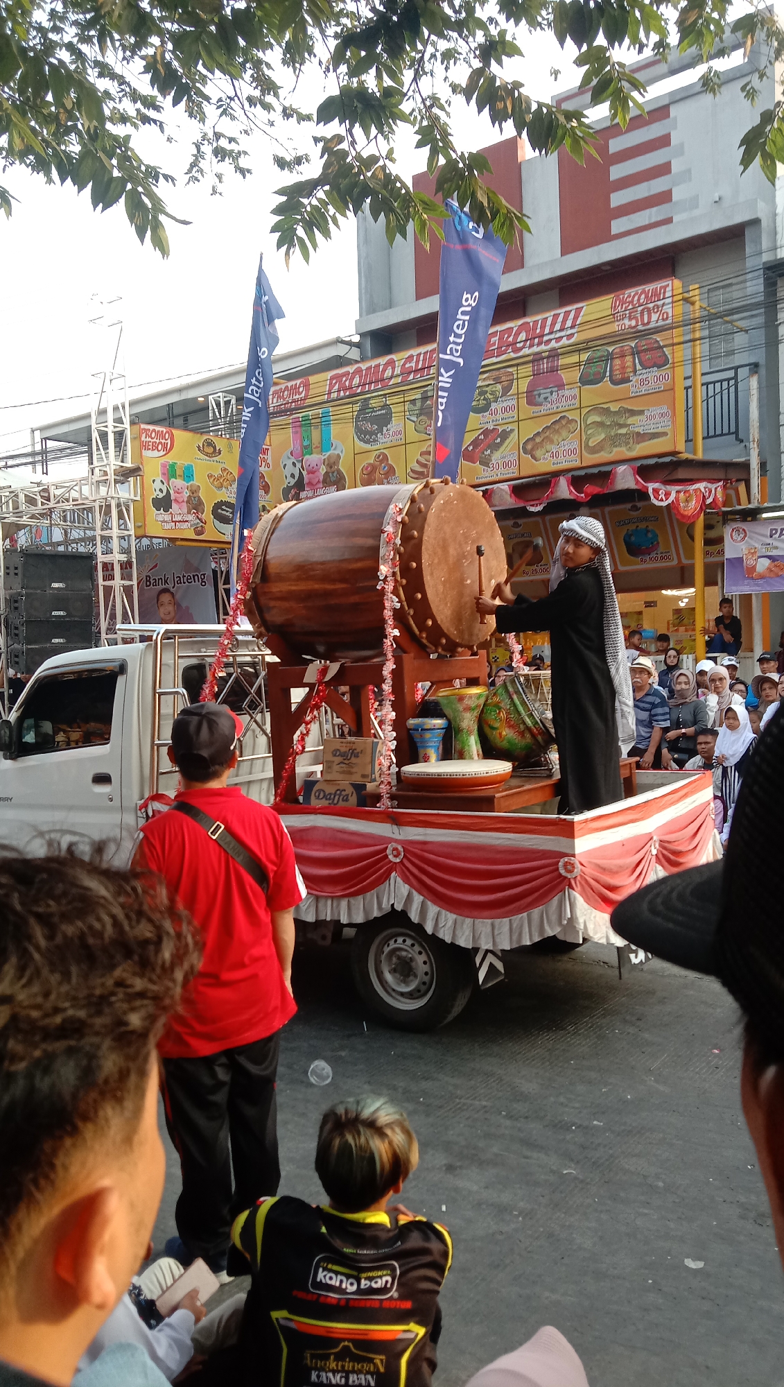 An outdoor daytime scene in Jatisawit, Indonesia, features a decorated truck as a stage. A young man wearing traditional Islamic attire, a thobe and a white and black checkered headscarf, is playing a large wooden drum with a mallet. He is positioned on the truck bed, which is adorned with red and white draped fabric and red streamers. In front of him are several smaller drums and other decorative items.
The truck is white and has the word "CARRY" visible on the side. Behind the truck, a building with a red and white striped facade is visible, along with banners advertising "PROMO SUPER HEBOH!!!!" and "Bank Jateng." A crowd of people are gathered, watching the performance, some standing and some seated. In the foreground, a person in a red shirt and black pants is facing away from the camera, also observing the event. Another individual in the foreground, seen from behind, wears a black jacket with yellow accents and the words "kang ban" visible on the back. The ground is paved with asphalt. The sky is visible above, with some trees and their leaves in the upper portion of the image.