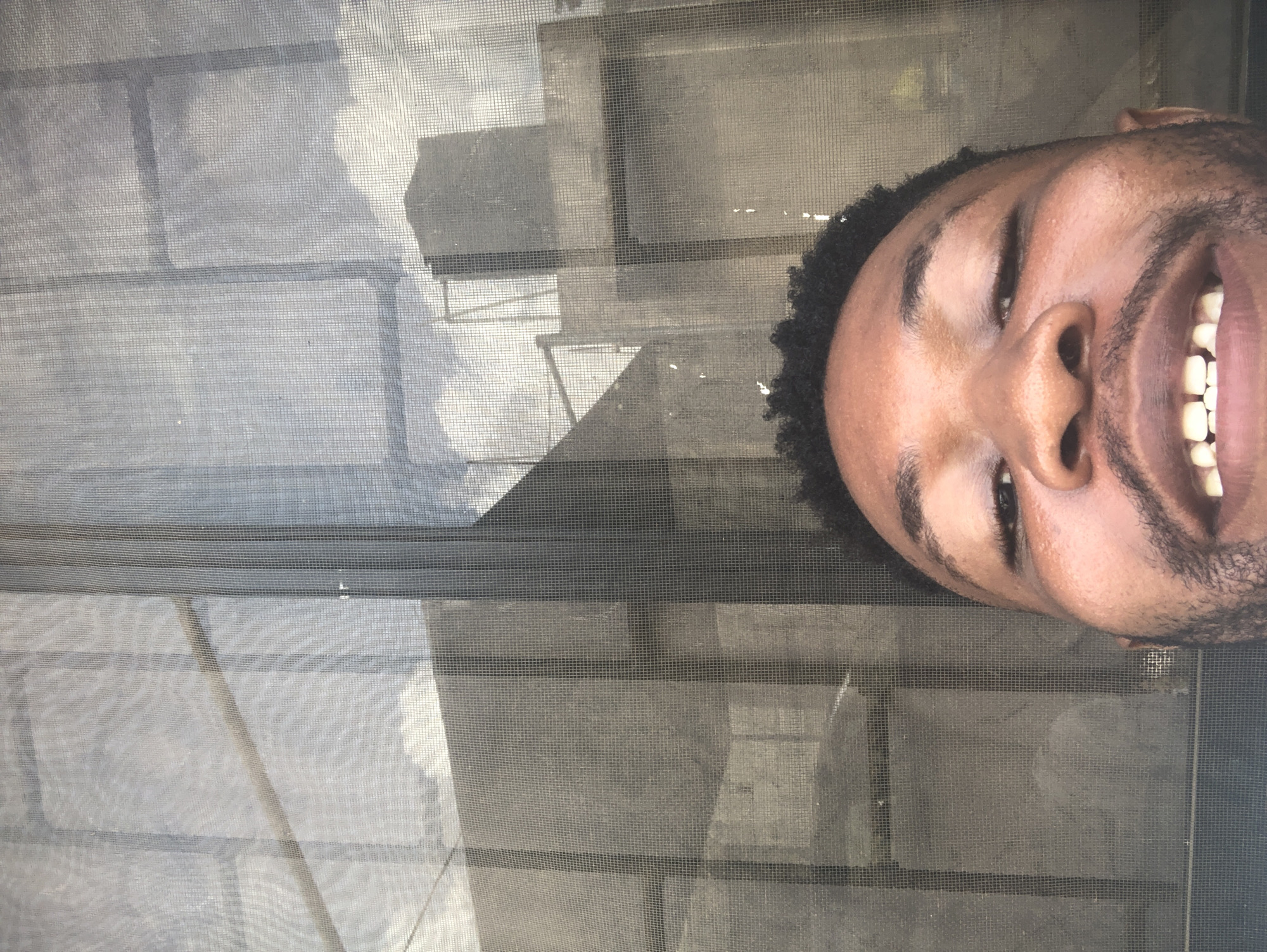 A medium shot looking through a window screen at a smiling man of African descent. His head is positioned to the right of the frame, with his face tilted upward. The background consists of a window and visible architectural elements through the screen. The location appears to be an urban environment, with window structures visible through the mesh.