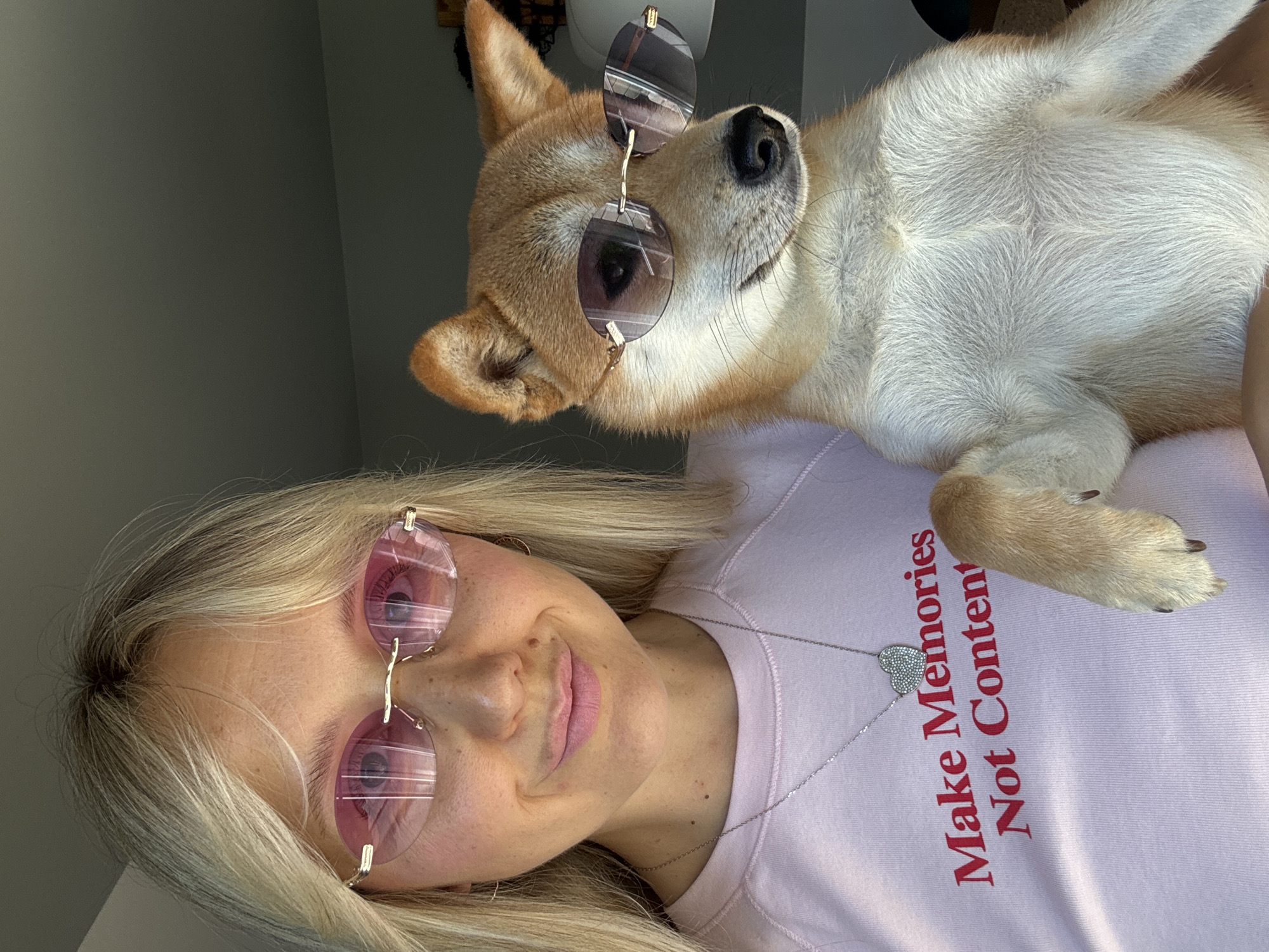 In a domestic indoor setting, possibly a home in Carnikava, Latvia, a blonde woman with fair skin and freckles, wearing a light pink sweatshirt with the text "Make Memories Not Content" in red and a silver heart pendant necklace, poses for a photo. She has on rimless pink-tinted sunglasses. To her right, and slightly in front, a tan and white Shiba Inu dog is held up, also wearing similar rimless pink-tinted sunglasses. The dog is looking to its right with its mouth slightly open. The background is blurred and appears to be a wall. The lighting suggests natural light coming from the left.
