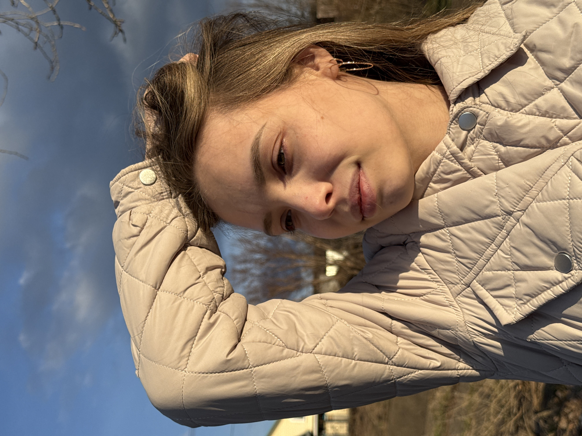 This close-up portrait, taken in Chaikovsky, Russia, features a young woman outdoors. Her head is tilted to her right (viewer's left), and she looks downwards with a gentle, subtle smile. She has long, light brown hair, and golden hoop earrings are visible in her left ear. She is wearing a light beige, diamond-quilted jacket with metallic snap buttons. One hand is partially visible, raised near her head, possibly running through her hair.
The setting is outdoors under a clear, bright blue sky with faint white clouds. The warm, direct sunlight illuminates her face and hair, indicating it is daytime, likely a sunny morning or afternoon. In the background, out-of-focus bare tree branches are visible against the sky, suggesting a colder season like late autumn or early spring. The ground, partially seen, shows dried grass or sparse vegetation. The overall mood is calm and serene. There is no visible text in the image.