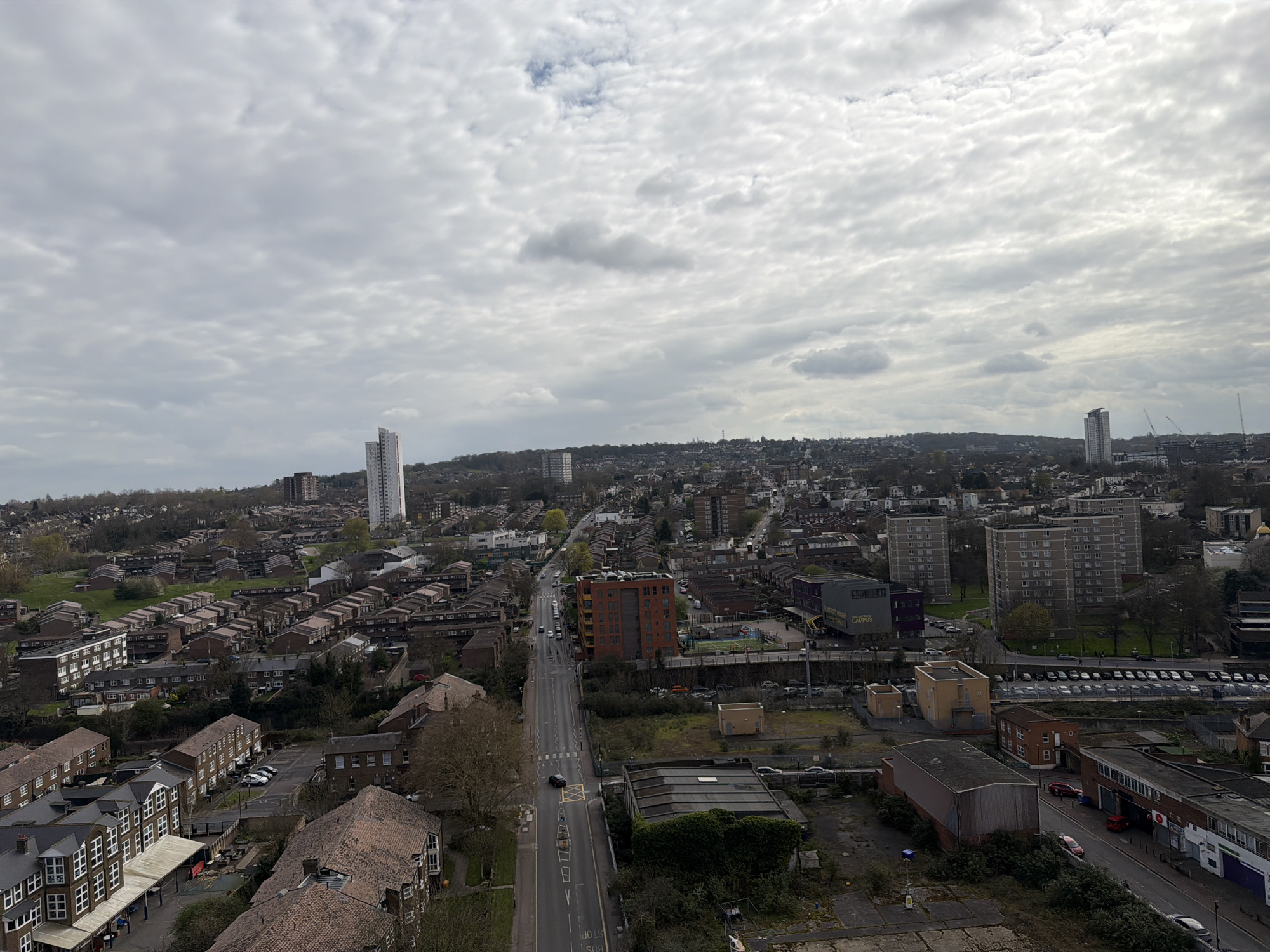The image shows an elevated view of a suburban neighborhood in London, United Kingdom, on an overcast day. A main road runs through the center of the frame, lined with buildings and trees. Residential areas with rows of houses are visible on both sides of the road. There are several high-rise apartment buildings scattered throughout the neighborhood, and cranes are seen on the horizon. The foreground shows industrial buildings, a large parking lot, and some greenery. The sky is filled with clouds.