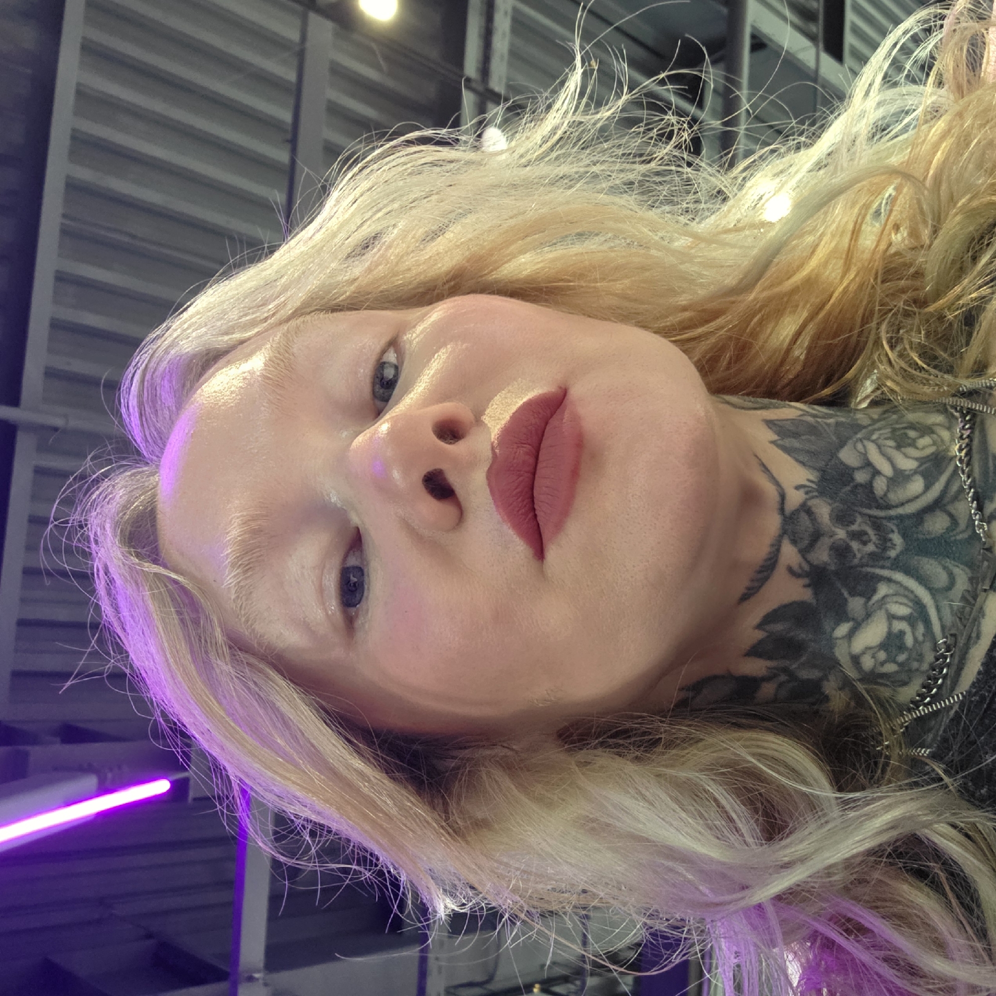 A close-up, angled portrait of a fair-skinned person with platinum blonde hair. The person has a neck tattoo and is wearing lipstick. A chain necklace is visible. The background features a corrugated metal ceiling and a blurred purple light source. The location may be in a building in Saint Petersburg, Russia, due to the person's possible cultural and ethnic background.