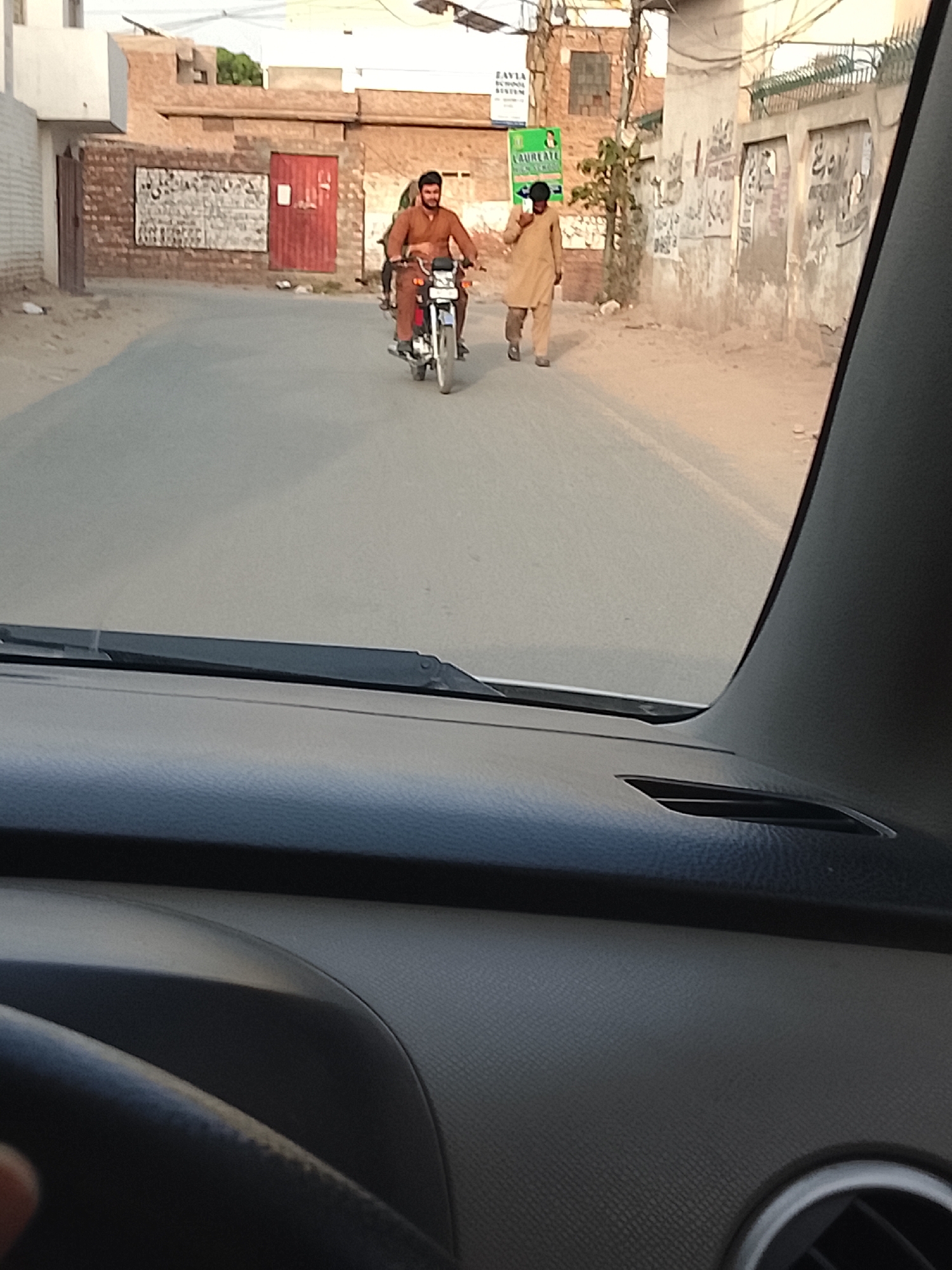 The image depicts a street in Nasheman Colony, Pakistan, viewed from inside a car. A man on a motorcycle is driving towards the camera. Another man in a traditional salwar kameez is walking beside him. Buildings line the street, including one with a sign for "ZAVIA SCHOOL SYSTEM." The street appears to be paved. The weather is sunny.