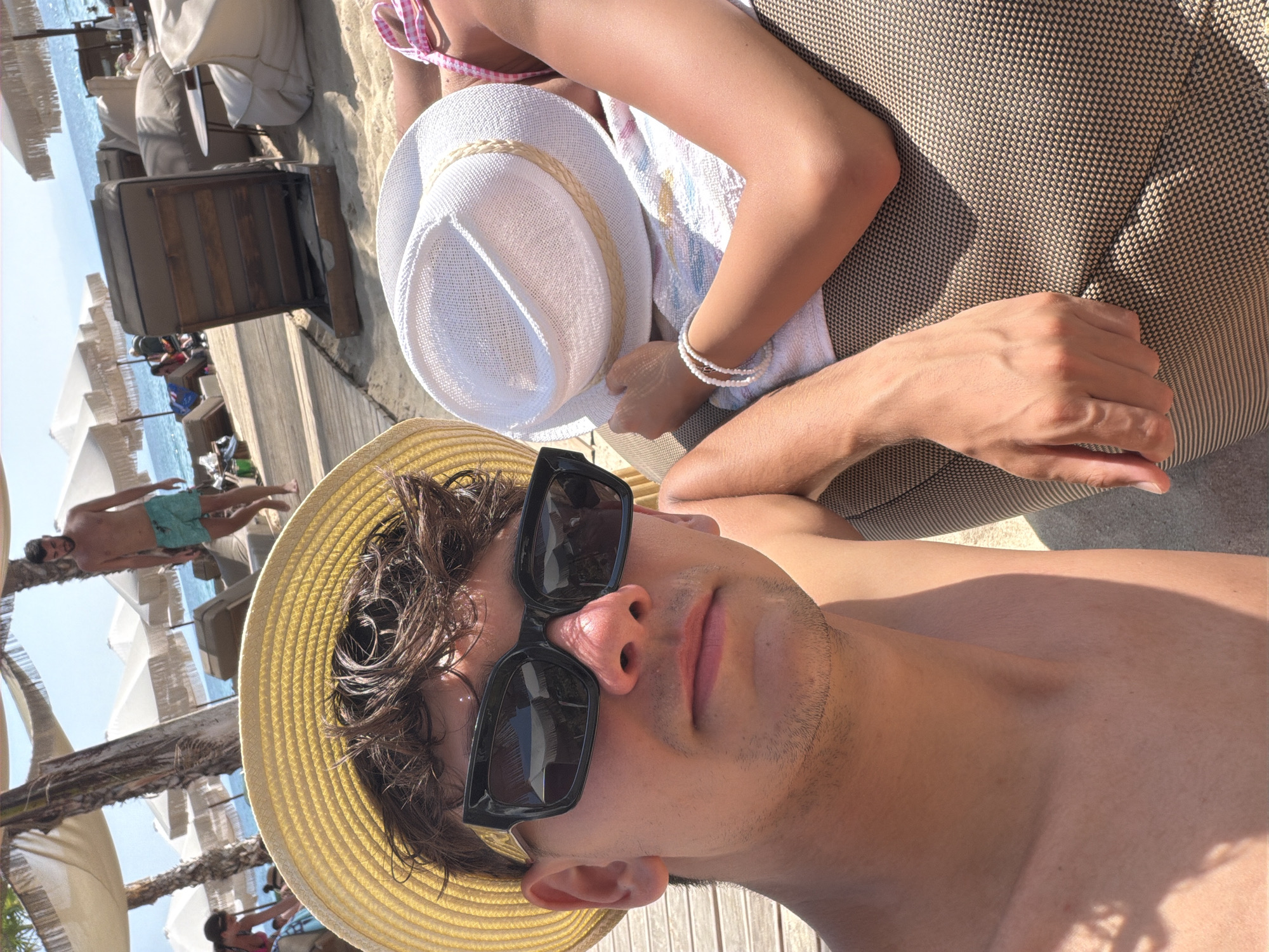A man wearing a yellow straw hat and sunglasses takes a selfie on a sunny day at Ofrynio Beach, Greece. He is shirtless and lying down. Next to him, another person wearing a white hat and a pink and white checkered swimsuit is also lying down. The background shows lounge chairs, umbrellas, palm trees, and the blue sea. In the distance, another man in blue swim trunks walks along the beach. The overall scene depicts a relaxed beach vacation atmosphere.