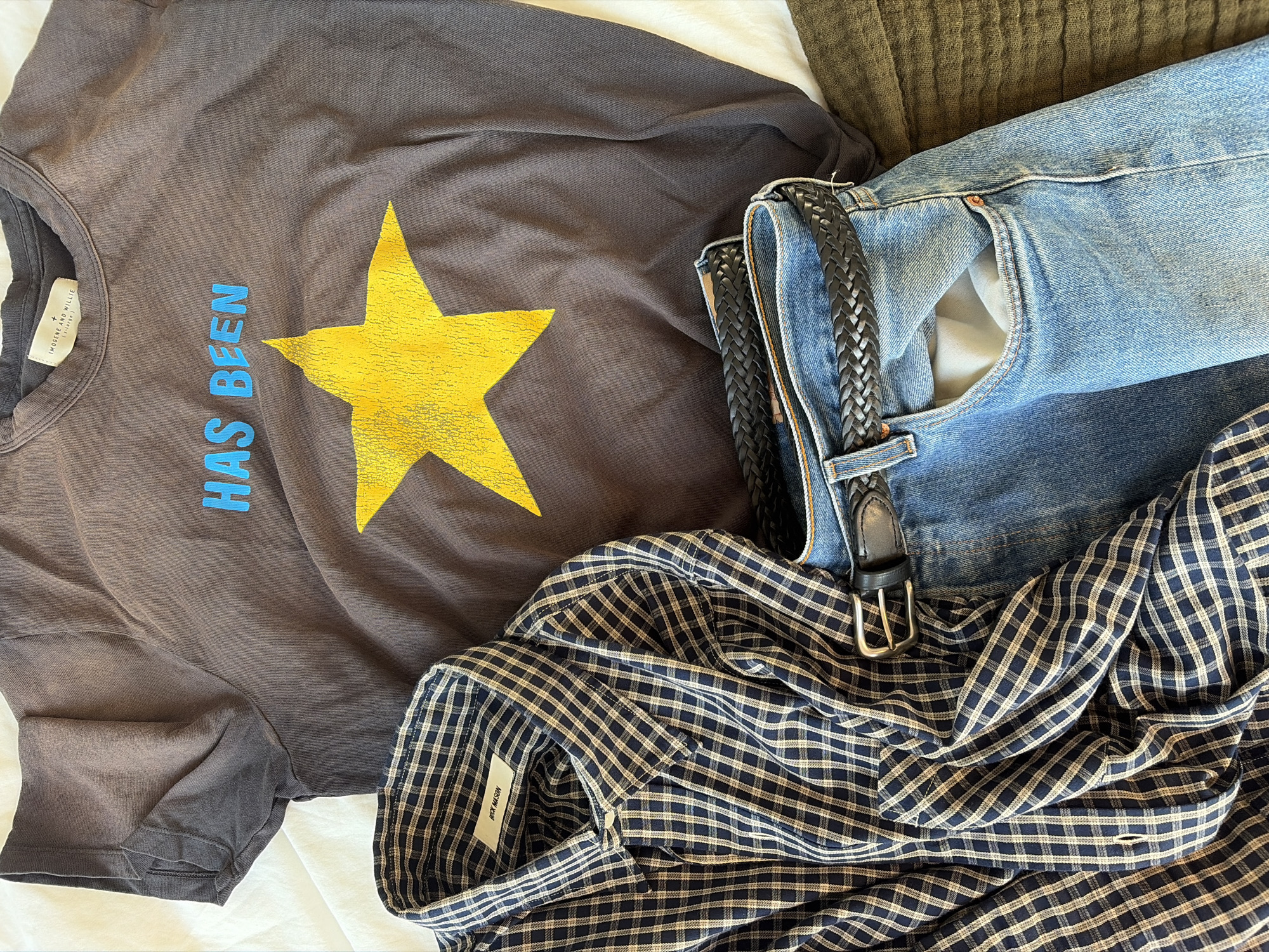 The image presents a flat lay arrangement of men's apparel on a light-colored, possibly white, fabric surface. Dominating the upper portion is a dark grey, short-sleeved t-shirt featuring the light blue text "HAS BEEN" centered above a large, distressed yellow five-point star graphic. A white label bearing the text "IMOGENE AND WILLIE" is visible on the t-shirt's interior collar. To the left and partially overlapping the t-shirt, a long-sleeved button-up shirt with a small blue and off-white checkered pattern is laid out; its collar displays a white label with "RICK MASON" printed on it. Below the t-shirt and partially beneath the checkered shirt are light blue denim jeans, through whose belt loops a black braided leather belt with a silver-toned rectangular buckle is threaded. A portion of olive-green fabric is visible in the bottom right corner, beneath the jeans. The scene depicts an indoor setting, and is located in the city of New York, United States of America.