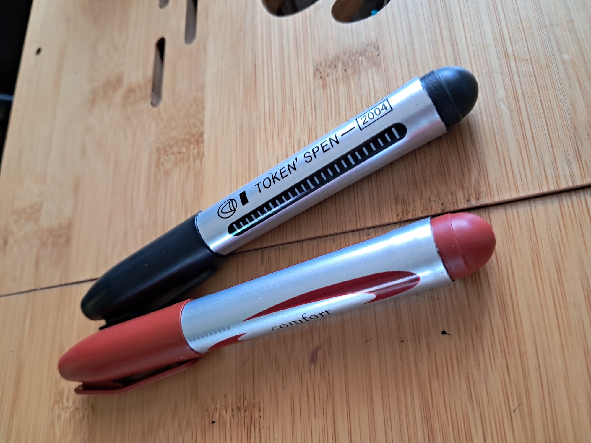 Two pens are lying on a light-colored wooden surface, possibly a desk or table, in an indoor setting in Kyiv, Ukraine. The wood has a distinct grain, suggestive of bamboo, and features several narrow, elongated cut-outs in the upper background.
The top pen has a silver barrel with a black cap and a black clip. Near its center, it features a logo resembling a stylized 'T' or 'D' and the text "TOKEN' SPEN — 2004". Below this text is a series of small, vertical etched lines. The end of this pen, which appears to be a stylus tip, is black and rounded.
The bottom pen, positioned slightly below and to the right of the first, also has a silver barrel but is accented with red. It features a red cap and a red clip. Along its barrel, a red swish design is visible, and the word "comfort" is printed in a script font. Its end, also a rounded stylus tip, is red.
The pens are simply resting on the surface, and there is no discernible activity taking place. The lighting suggests ambient indoor illumination. No specific time of day or weather conditions can be determined from the image.