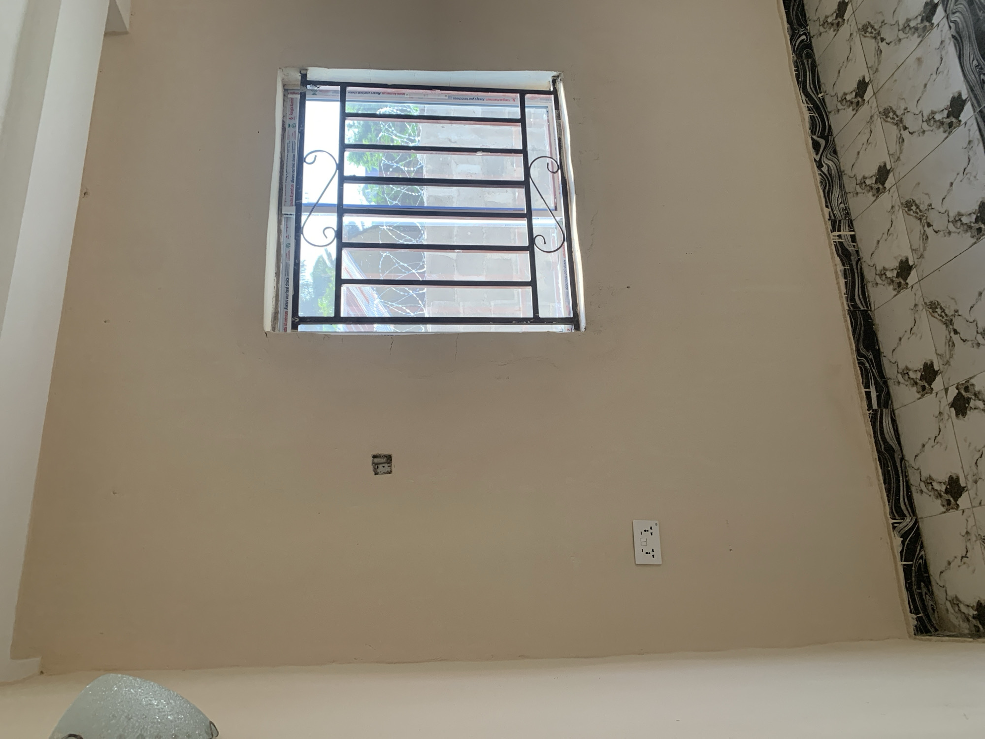 The image shows an interior view of a room with light-colored walls and tiled flooring. A window with black metal bars is present on the wall, with barbed wire visible outside. Electrical outlets are installed on the wall, one of which appears to be a double socket. The floor is covered with marble-patterned tiles. In the upper left corner, a light fixture is partially visible. The environment appears to be a residential or commercial space under construction or renovation, given the unfinished appearance of the walls and the presence of exposed electrical fittings. The location is identified as Unknown, Unknown.