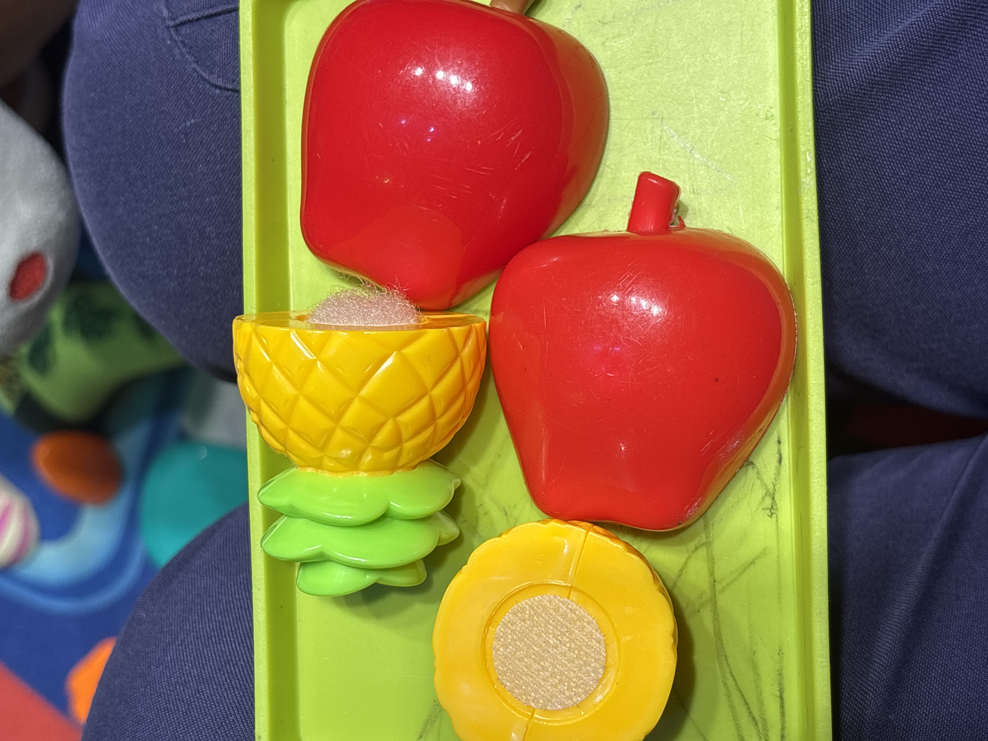 A close-up, overhead shot showcases a bright green plastic tray containing several colorful toy fruits. On the left, a sliced yellow toy pineapple is visible, with its green leafy crown detached and placed beside it. Next to the pineapple, on the left side of the tray, is a yellow toy object with a circular, textured brown center, resembling a cross-section of a vegetable or fruit. The right side of the tray holds two bright red toy objects that appear to be bell peppers. The toy pineapple has a rough, textured surface, and its cut surface reveals a hook-and-loop fastener. The toy peppers have a smooth, glossy finish and one of them has a small red stem-like protrusion. The tray itself has faint, dark green scribbled lines on its surface. The background is out of focus but suggests a colorful, possibly child-oriented environment with blurred shapes of blue, orange, and green. The tray is placed on top of dark blue fabric, which is likely clothing. There is no indication of the location being Dubai, United Arab Emirates; it is a generic indoor setting.