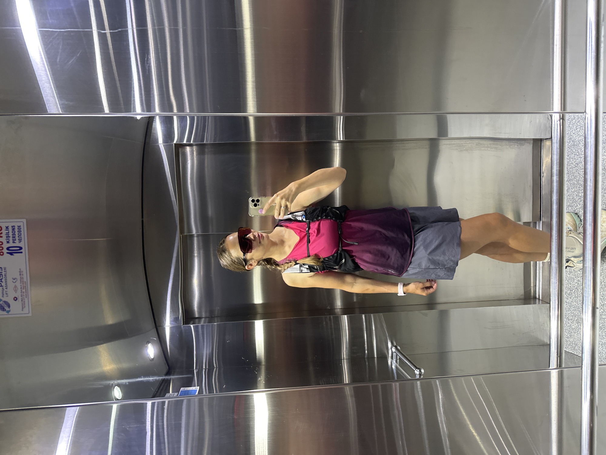 A female stands in the corner of a metallic elevator taking a selfie with a smartphone. She wears sunglasses, a pink and purple tank top, grey athletic shorts, a black backpack, and a white wrist band. The elevator's interior is stainless steel, and there are metal railings near the door. A sign on the back wall indicates a maximum capacity of 10 people and 800 kg. The sign includes Turkish text and the words "Oasis" and "Lift Asansörleri." It is likely located in Fethiye, Turkey based on the textual information.