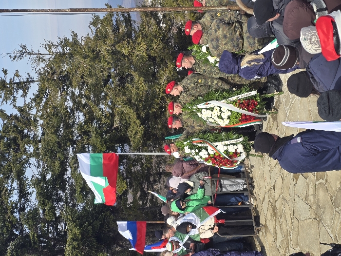 The image displays an outdoor ceremonial gathering on a stone-paved plaza, situated in the city of Shipka, Bulgaria. A large Bulgarian flag, white, green, and red, flies from a flagpole, with a smaller Russian flag visible on another pole to its left. The background consists of dense evergreen trees beneath a light blue sky. In the mid-ground, a line of approximately six individuals in green camouflage uniforms, red berets, and black boots stand holding large, ornate wreaths composed of evergreen foliage and white and red flowers with ribbon sashes. To their left, behind a low metal barrier, a group of civilians, including adults and children, observe the proceedings; several are draped in or hold small Bulgarian flags. The immediate foreground shows the backs of numerous attendees, clad in winter clothing, some holding small Bulgarian flags. The assembly appears to be participating in a commemorative or memorial event.