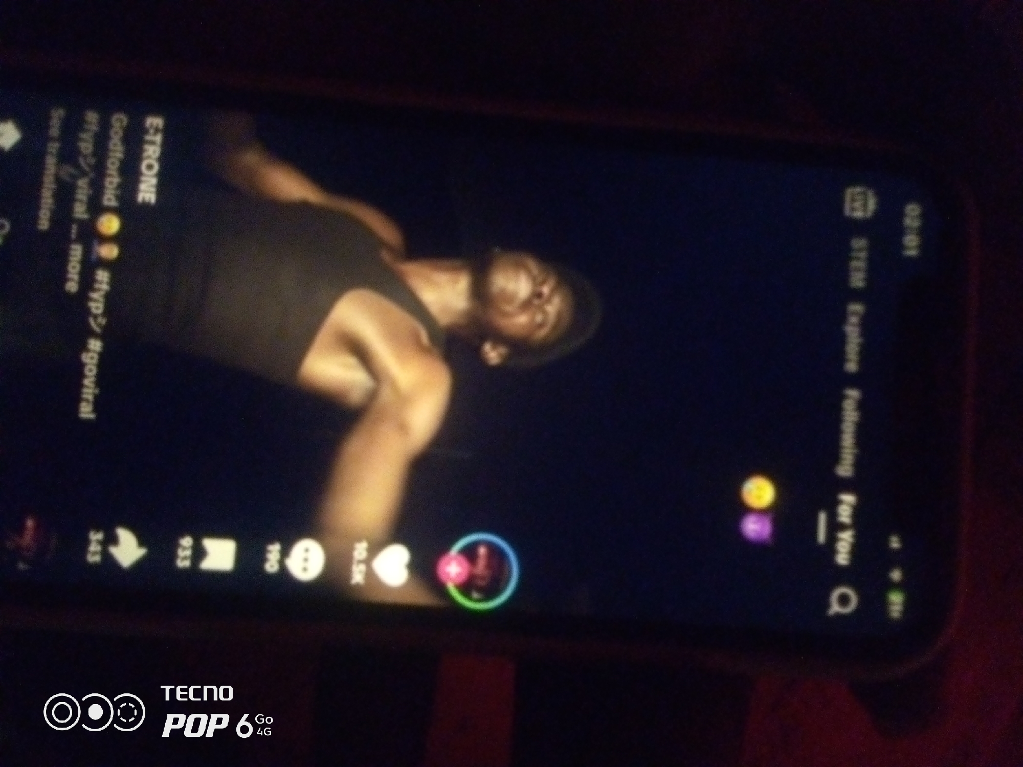 The image shows a close-up of a smartphone screen displaying a video on a social media platform, likely TikTok, given the interface elements such as hashtags, likes, comments, and shares. The video features a person, appearing to be a young man, wearing a dark tank top and looking upwards, with his face illuminated. The background of the video is dark. On the left side of the screen, there is text including "ETRONE," "Godforbid," and hashtags like "#fypシ," "#viral," and "#goviral." The bottom of the phone screen displays the "TECNO POP 6 4G" branding. The surrounding environment is dark, suggesting the photograph was taken in low light conditions. The image is not of sufficient detail or context to determine the exact location in Howayi, Nigeria.