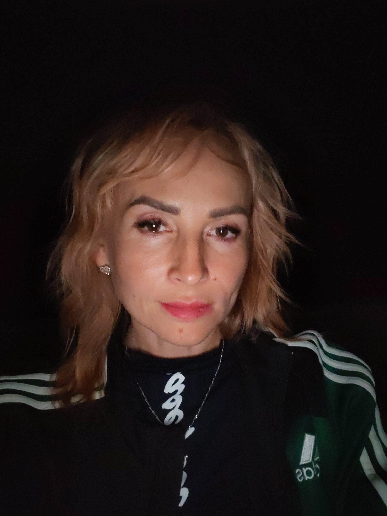 A woman with blonde hair and light skin is pictured in a close-up, selfie-style shot. She is wearing a black jacket with green and white stripes on the sleeves. The background is dark, suggesting an indoor or nighttime setting. The woman has on makeup, including pink lipstick and eyeshadow. She is also wearing a silver necklace and a small heart-shaped earring. The image appears to be taken in Ivanov Bor, Russia.