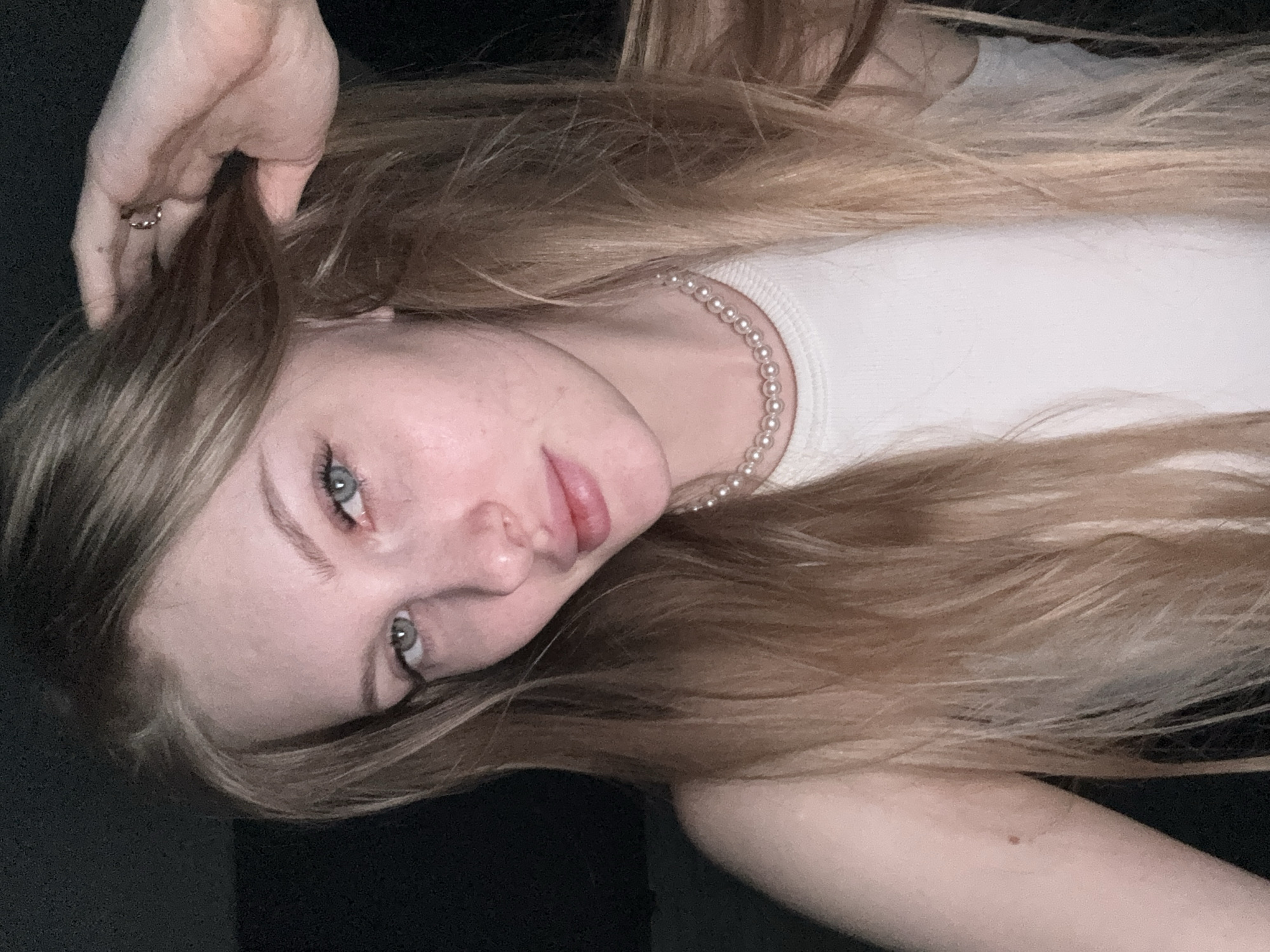 This media file is a close-up, rotated portrait of a young woman from Perm, Russia, looking directly at the camera. She has long, wavy hair, predominantly light blonde or golden-brown, framing her face. Her striking eyes are a clear blue-green, and her lips are full with a natural pink hue, slightly parted.
She is wearing a simple white top with a ribbed texture and a pearl necklace consisting of small, uniformly sized white pearls. Her right hand is raised to the side of her head, with her fingers gently touching her hair near her temple. A small, dark-toned ring with a subtle detail is visible on her ring finger.
The background is dark and appears to be a solid, indistinct surface, suggesting an indoor setting with focused lighting on the subject. There are no discernible location cues within the image itself, beyond the provided context of Perm, Russia. The time of day appears to be evening or night, given the dark background and apparent artificial lighting. Her expression is direct and somewhat serene or thoughtful. No visible text is present in the image.