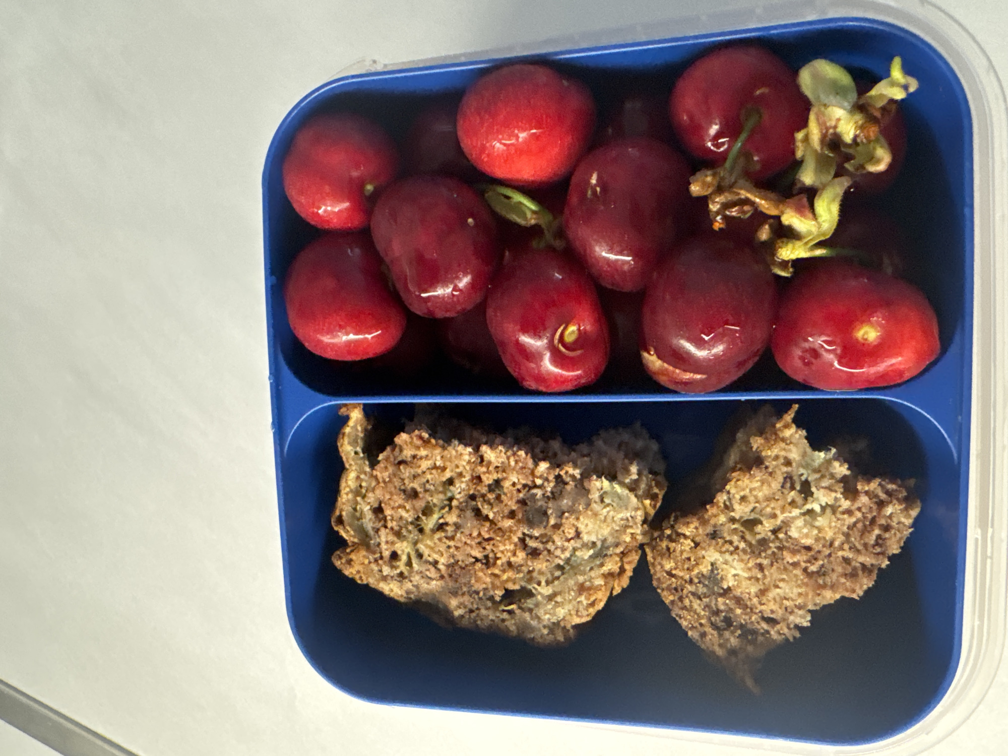 A blue, divided lunchbox sits on a white surface. The top compartment contains a cluster of dark red cherries, some with stems still attached. The bottom compartment holds two pieces of brown, textured bread, possibly zucchini bread or a similar type of loaf. The scene is devoid of any indication of location within Sofia, Bulgaria, or any human presence. The focus is solely on the contents of the lunchbox.