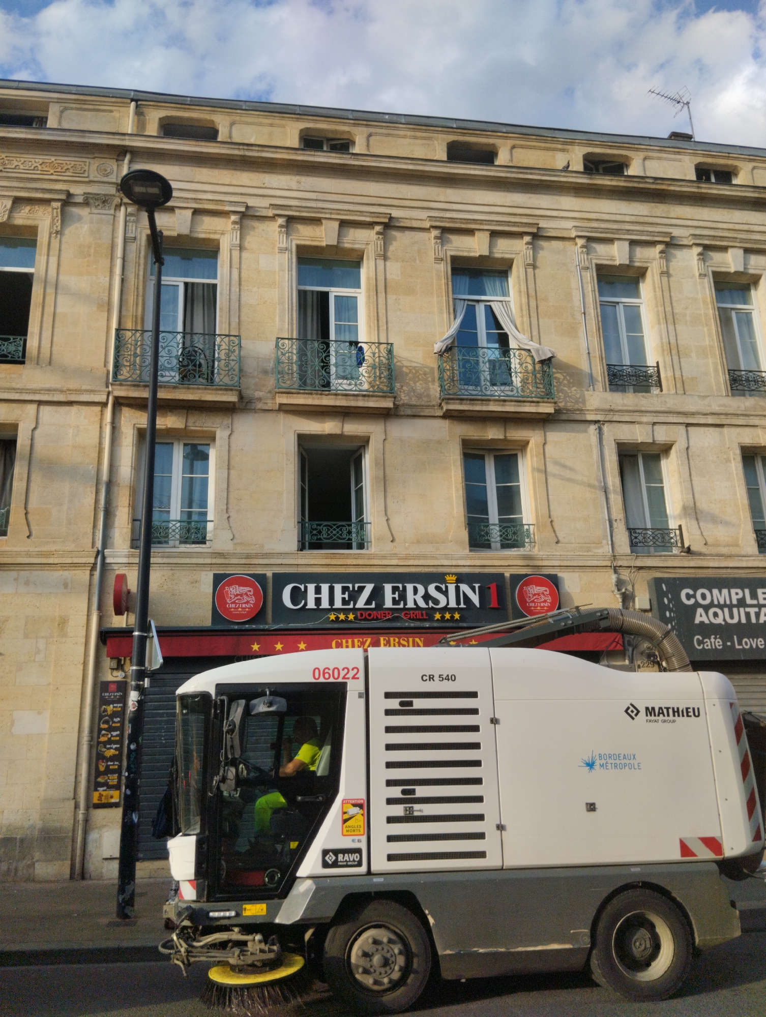 The image depicts a street-level view of a white street sweeper vehicle with the brand name "RAVO" visible on its side. The sweeper is positioned on a paved street in front of a multi-story building with light-colored stone facade and multiple windows. Above the building's ground floor, a red awning stretches across the facade, and signage indicates "CHEZ ERSIN 1" and "COMPLET AQUIT". The ground floor also has a menu board visible on the left side. A person in a bright yellow shirt and green pants is operating the street sweeper. The sky above is partly cloudy. The scene is an urban setting in Bordeaux, France, indicated by the "BORDEAUX METROPOLE" logo on the sweeper.