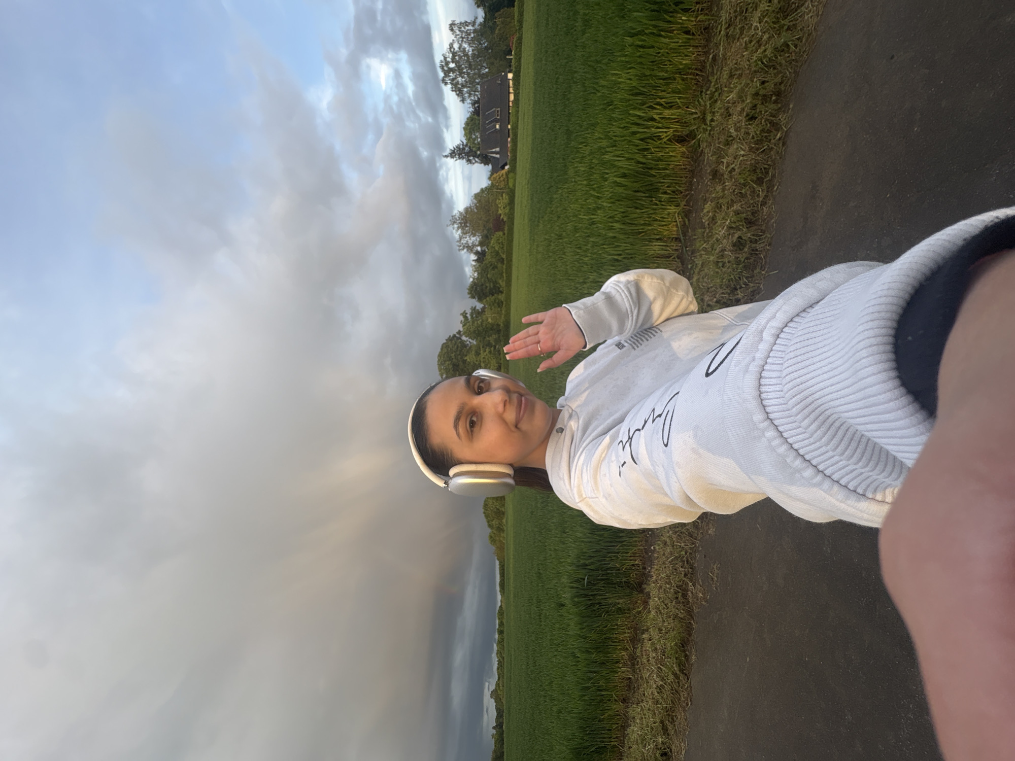 A woman is taking a selfie outdoors. She is wearing headphones, a white hooded sweatshirt, and dark shorts. She is on a paved path next to a field of green grass. In the background, there are trees and a house. The sky is cloudy, and a rainbow is faintly visible on the left side of the image. The photo was likely taken in a rural or park area near Mönchengladbach, Germany.