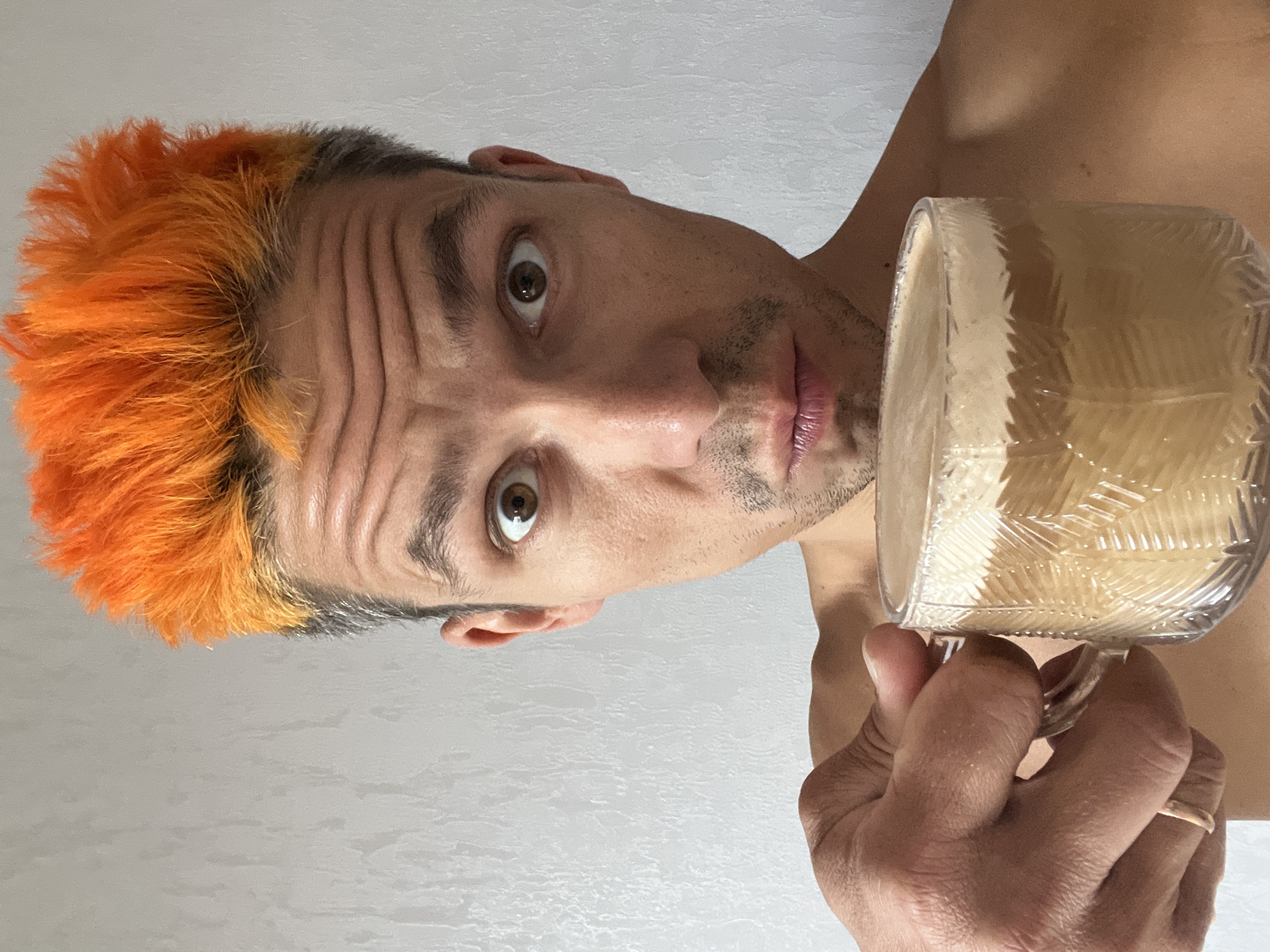A close-up, chest-up selfie of a man with bright orange hair and a surprised expression, holding a clear glass mug filled with a frothy beverage. He is shirtless, with his left hand holding the mug, and his right arm and shoulder are visible. The background is a plain, light-colored textured wall. The man is positioned in the center of the frame, with the mug prominently displayed in the foreground. The lighting is soft and even, highlighting the texture of the wall and the foam on the drink.