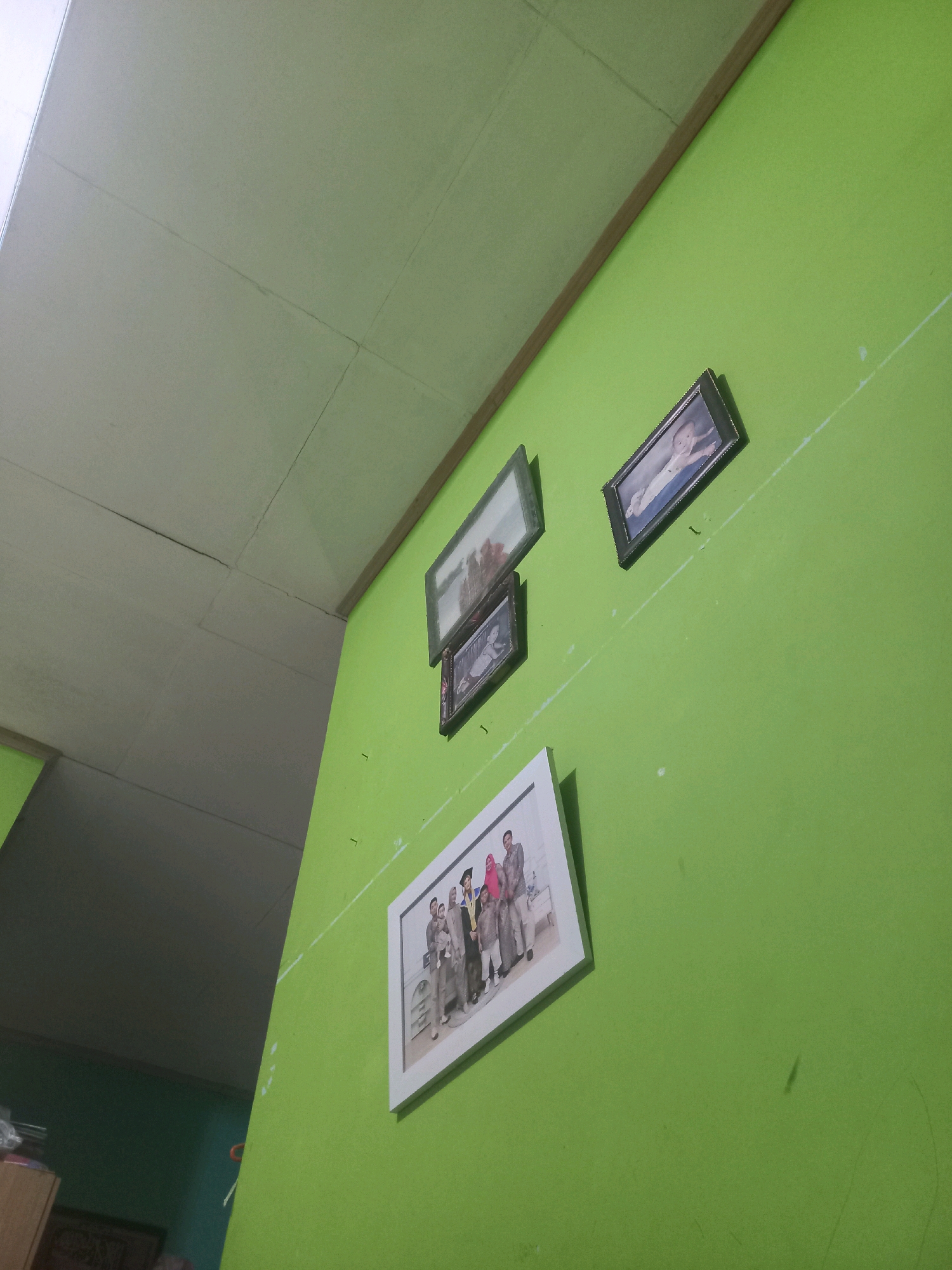 The image captures an interior view of a room with lime green walls and a white ceiling. Several framed photographs are displayed on the wall. In the upper right is a framed photo of a baby. Below and to the left of it, another framed photo shows a group of people, some wearing formal attire, including what appears to be graduation robes and head coverings. To the left of that, a smaller framed photo displays a landscape scene with trees. The ceiling is paneled with visible seams. The overall impression is of a domestic setting, possibly a living room or hallway, in a residence. The scene lacks any specific architectural or urban context to pinpoint a precise location within Sukoharjo, Indonesia, beyond being an indoor environment.