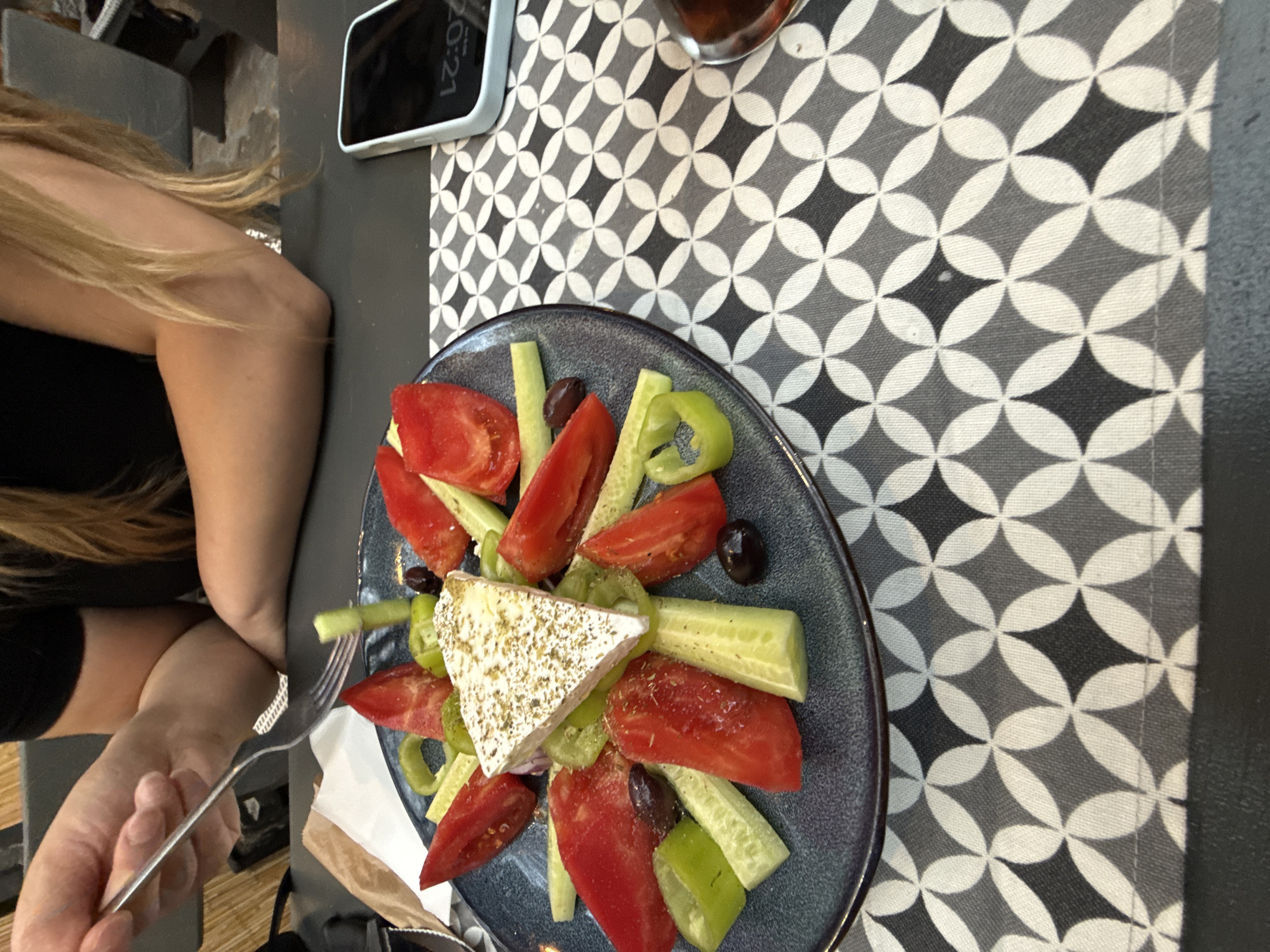 The image depicts a close-up overhead view of a Greek salad on a dark-colored plate. The salad contains large wedges of red tomatoes, cucumber spears, green bell pepper rings, black olives, and a triangular block of feta cheese sprinkled with herbs. The plate is set on a black table covered with a grey and white patterned tablecloth. A person's arm and hand, holding a fork, are visible on the left side of the frame, reaching towards the salad. In the background, another person's arm and shoulder, wearing a black top, are visible. A smartphone with a blank screen is placed on the table to the right of the salad, next to a glass of dark liquid. The scene appears to be at a restaurant. There is no information within the image or its context that indicates the location in the city of Stara Zagora, Bulgaria.