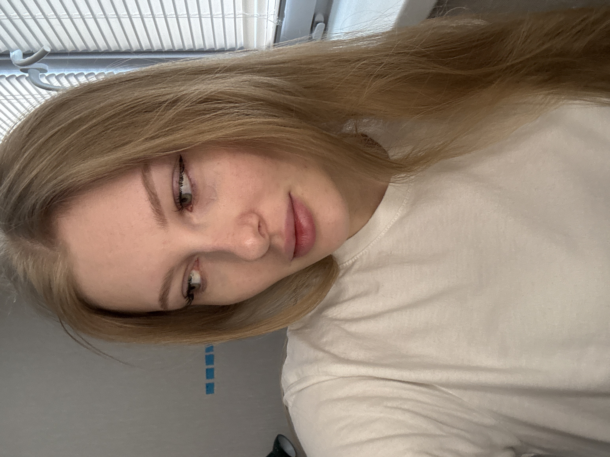 A selfie photograph shows a young woman with long blonde hair wearing a white t-shirt. She is inside, likely near a window with blinds. Three small blue squares are visible on the wall behind her. The photograph was probably taken indoors, possibly in Perm, Russia.
