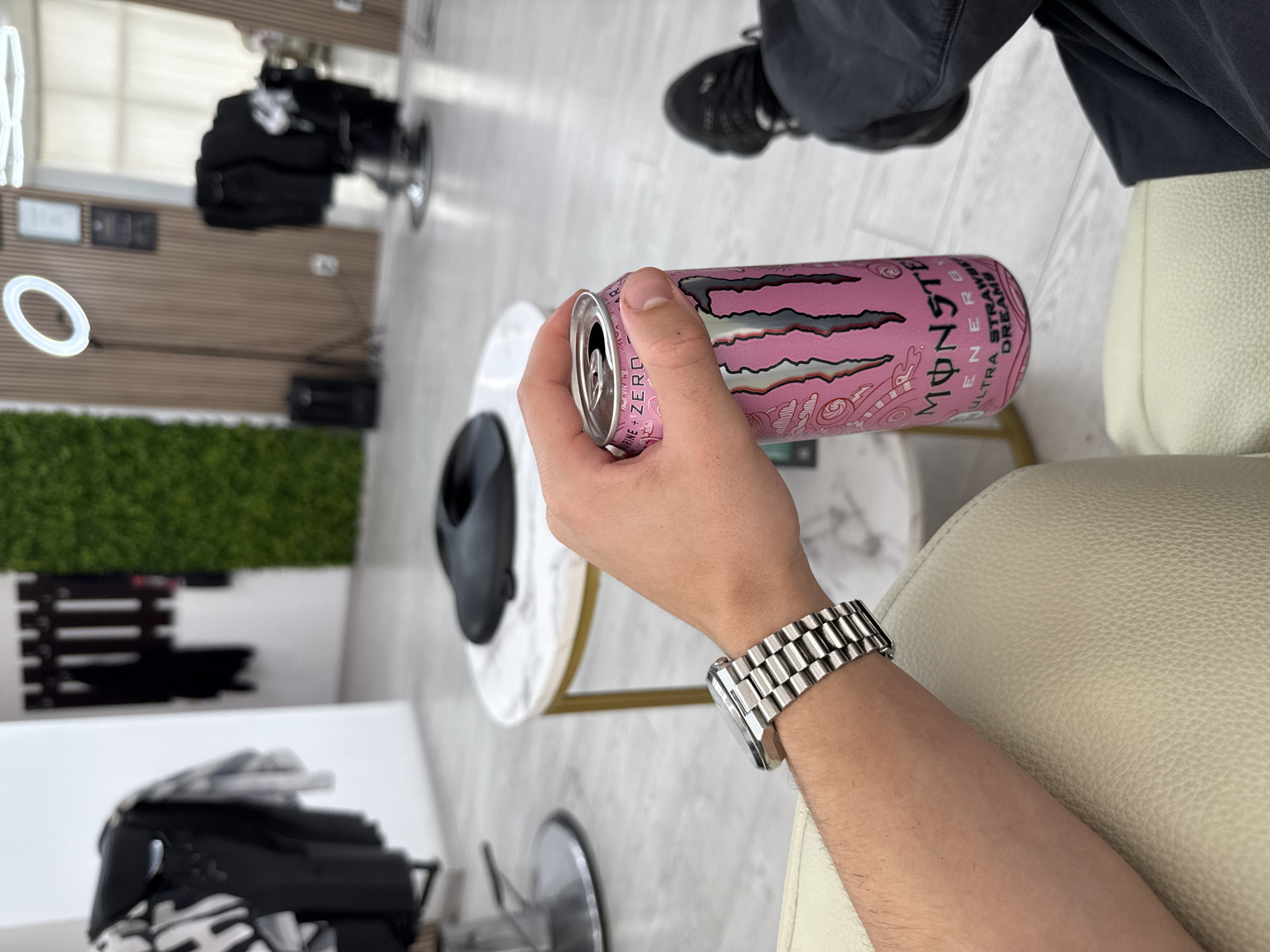 A person is holding a pink Monster Ultra Strawberry Dreams energy drink can in their right hand, wearing a silver wristwatch. The person appears to be seated on a beige leather chair, with their legs visible, wearing dark grey trousers and black sneakers. A small, round white table with a gold frame is next to them, displaying a black hat. The background features a salon or barbershop environment, with styling chairs, mirrors, and green decorative plants. The location is likely in the city of Sofia, Bulgaria.