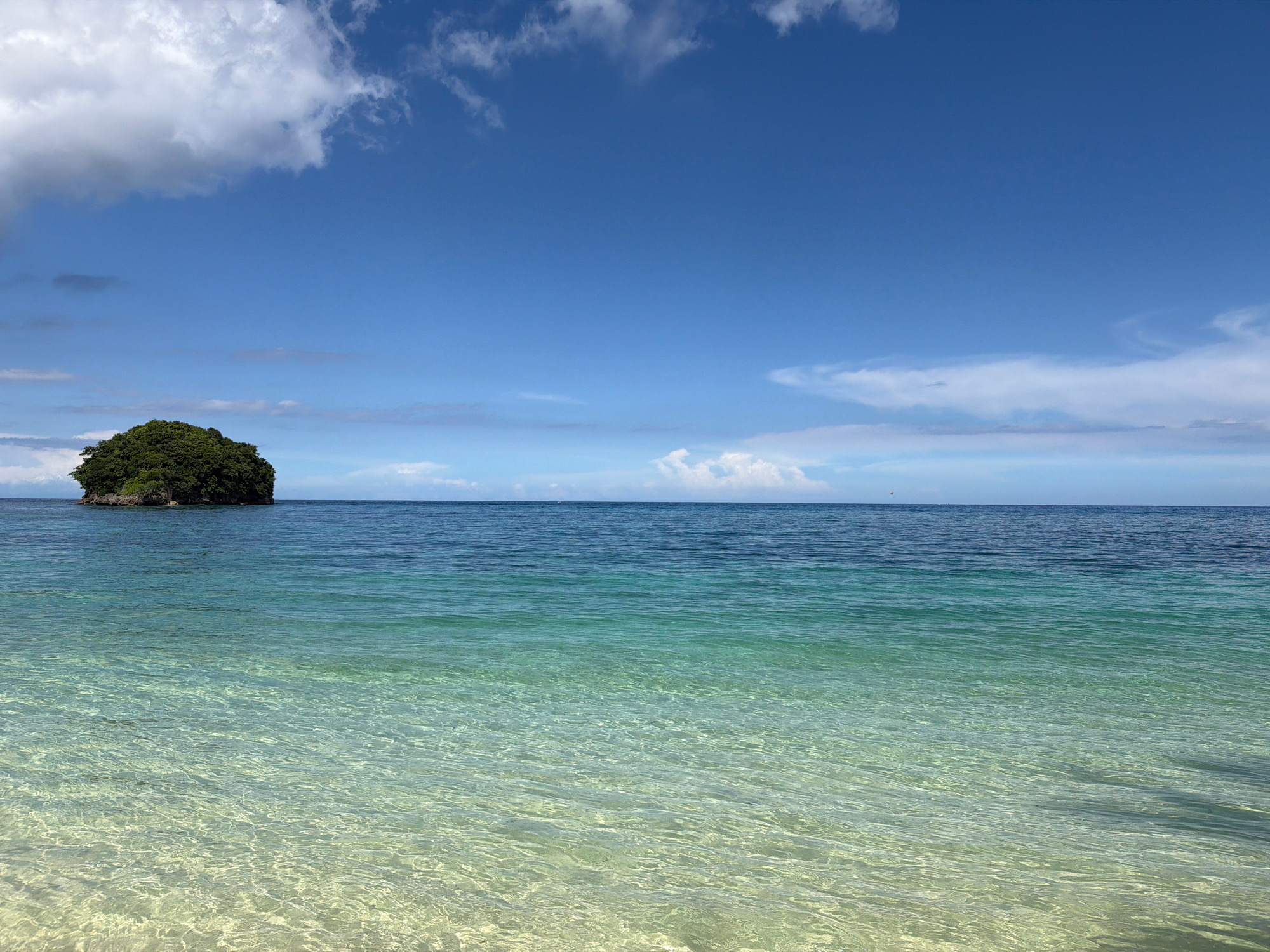 The image depicts a tropical seascape with a clear blue sky filled with scattered white clouds. The ocean stretches across the frame, transitioning from a vibrant turquoise near the shore to a deeper blue horizon. In the distance, a small, lush green island rises from the water, adding a solitary focal point to the expansive sea. The foreground features shallow, crystal-clear water, revealing the sandy seabed below with gentle ripples distorting the light. This scene is typical of the coastal areas in Malay, Philippines, known for its pristine beaches and islands.