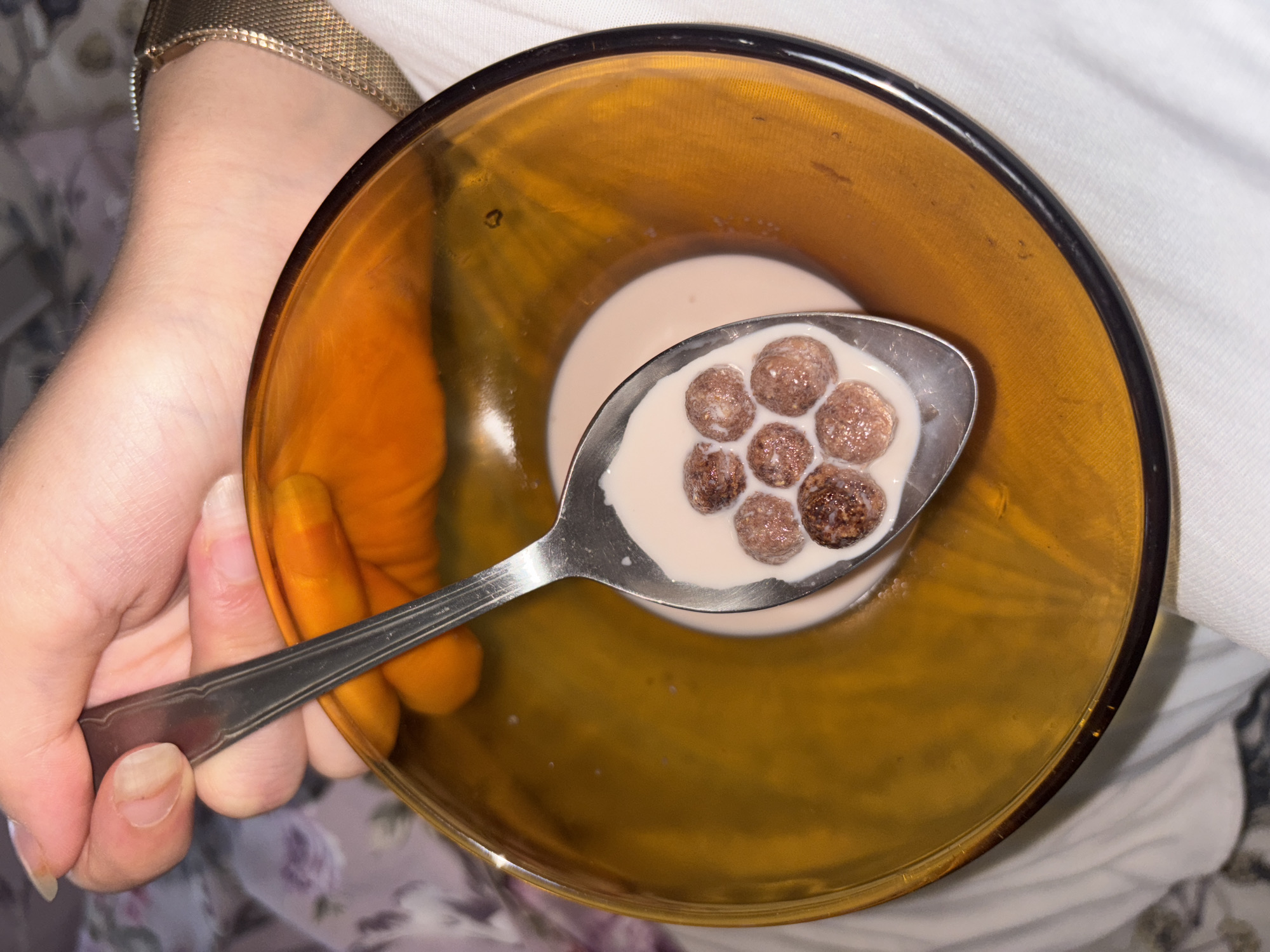 A person's hand is holding a metal spoon filled with chocolate cereal and milk over a translucent amber bowl containing milk and cereal. The bowl is partially filled with milk, and a few pieces of chocolate cereal are visible floating on the surface. The spoon is angled downwards, scooping up some of the cereal and milk. The person's hand is positioned over the bowl, with fingers wrapped around the spoon's handle. A wrist watch with a metallic band is visible on the person's wrist. The background is softly blurred, suggesting an indoor setting with patterned fabric and white material. There is no information available to determine the location in Vratsa, Bulgaria.