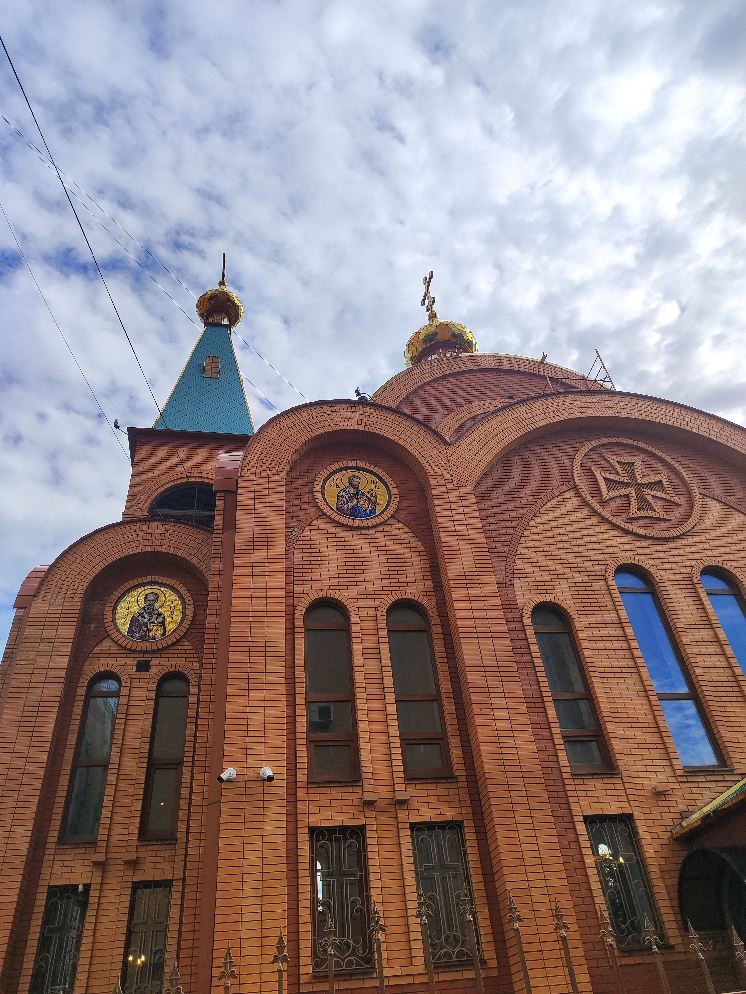 An upward-angled shot captures the imposing facade of an Orthodox church in Krasnodar, Russia, under an overcast sky. The building is constructed primarily of reddish-brown brick, featuring intricate arched designs and patterns.
Two golden, onion-shaped domes are visible towards the top. The one on the left sits atop a conical, blue-teal colored roof section, while the larger dome on the right crowns the main structure. Both domes are surmounted by traditional Orthodox crosses.
The facade is adorned with circular elements. On the left side, a mosaic depicts a bearded saint, likely holding a book, with faint Cyrillic text above his head. On the right, another circular mosaic features a different bearded saint, clad in religious vestments, with the Cyrillic inscription "СВ. НИКОЛАЙ" (Saint Nicholas) partially visible above. A large circular brick relief of an Orthodox cross is prominently displayed on the upper right section of the facade.
Multiple arched and rectangular windows punctuate the brickwork; some reflect the cloudy sky, while others on the lower levels are protected by ornate metal grates with cross patterns. Two white security cameras are mounted on the brick wall below the left mosaic. Faint power lines stretch across the cloudy sky in the upper left corner.
No people are visible, and no specific activity is occurring, but the church appears to be a well-maintained place of worship. The overall impression is one of spiritual architecture set against a dramatic sky on what appears to be a cloudy daytime.
