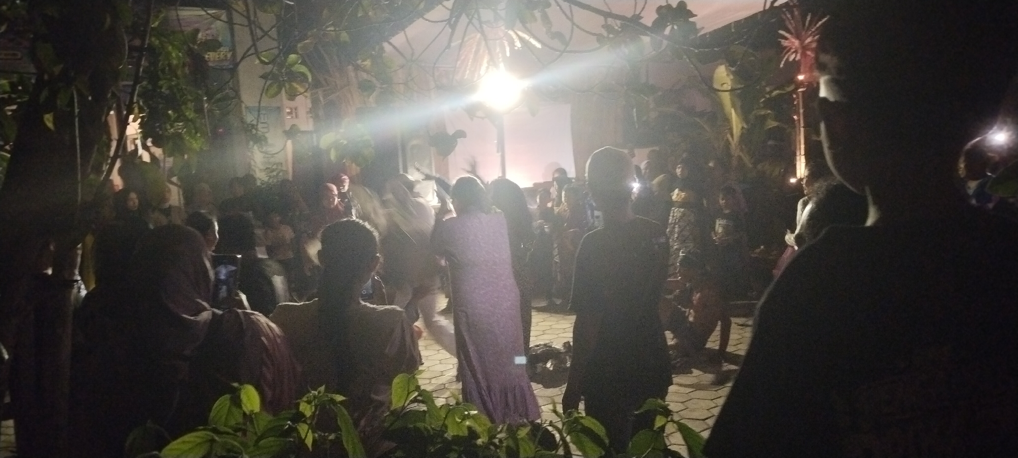 The image depicts a nighttime gathering of people in an outdoor setting, possibly Keting, Indonesia. The scene is illuminated by artificial lights, creating a somewhat hazy atmosphere. A large crowd is visible, with individuals standing and observing. In the foreground, a person's back is to the camera, obscuring their face. To their left, another individual is partially visible, also with their back turned. A woman in a purple, patterned dress is standing in the mid-ground, facing away from the viewer and towards the crowd. The background shows more people, some holding what appear to be mobile phones, suggesting they are documenting the event. Overhead, there are branches and foliage, suggesting the event is taking place in an area with trees or natural elements. A bright light source, possibly a spotlight or strong lamp, is visible in the upper center of the image, casting a glow. In the upper right, a vertical structure with what looks like a fireworks display or a decorative light fixture is present. The ground appears to be paved with interlocking stones. The overall impression is of a community event or celebration occurring after dark.
