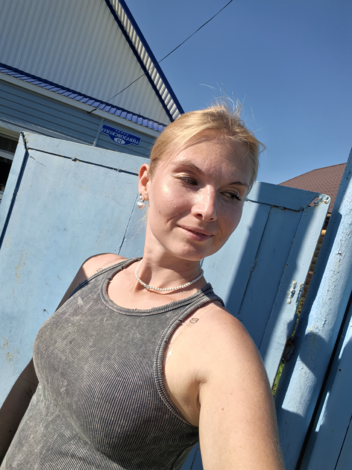 A woman with blonde hair and fair skin, wearing a gray ribbed tank top and a pearl necklace, is photographed from the chest up outdoors. She has a slight smile and is looking to her right. A blue wooden gate and a blue building are visible behind her, along with a clear blue sky. A street sign with Russian text is mounted on the building, identifying the location as Sterlitamak, Russia, specifically on Oktyabrskaya Street (ул. Октябрьская). The lighting suggests it is a sunny day.